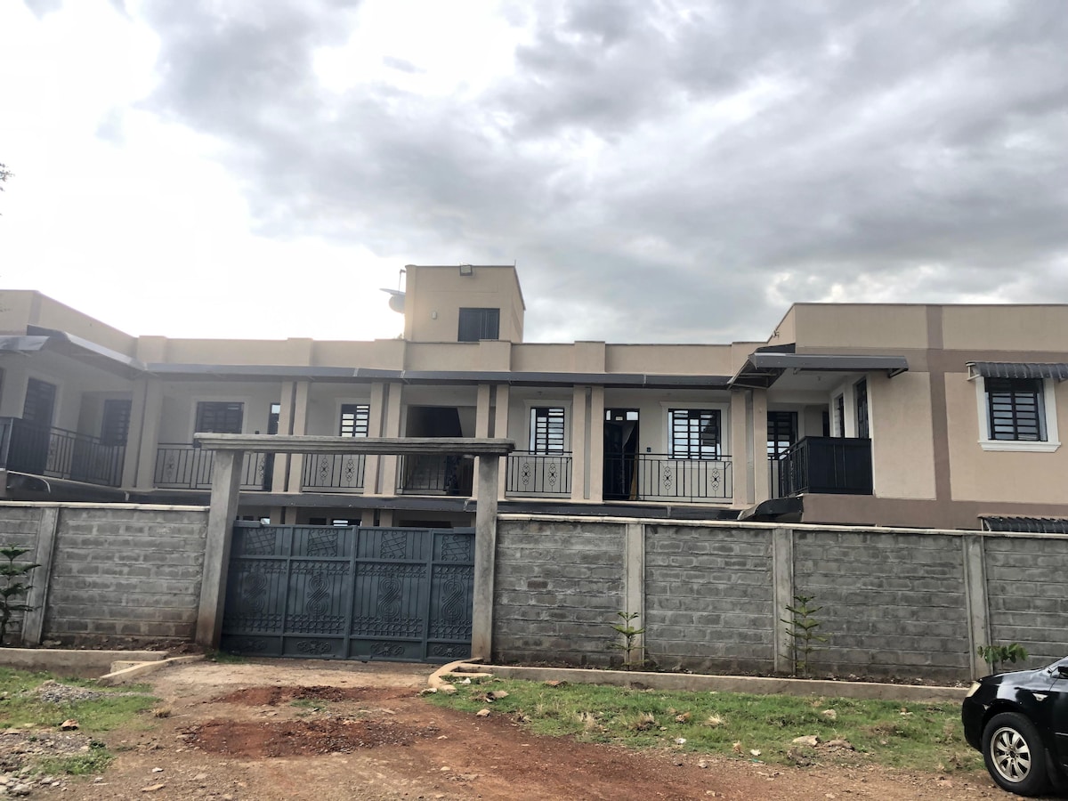 The exterior of the building is showcased, featuring a two-story design with multiple balconies and large windows. A secure gate is visible, leading to the entrance. A gravel area is present in front, with a blend of grass and shrubs along the perimeter.