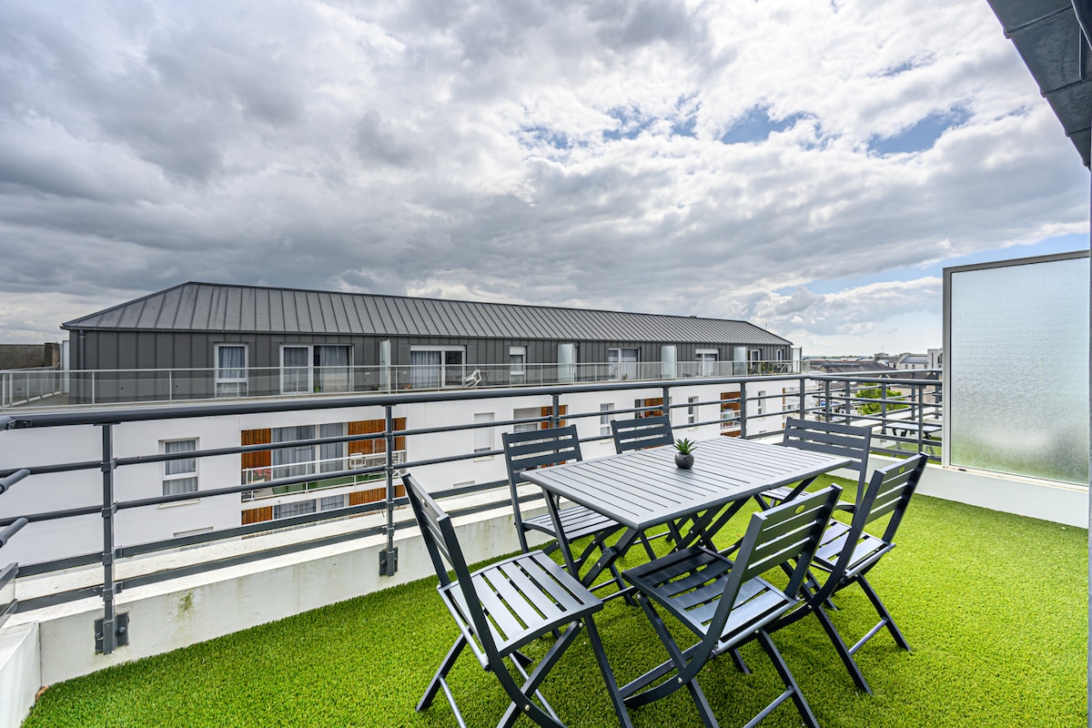 A private terrace features a table and several chairs, set against a backdrop of rooftops under a partly cloudy sky. The space is enhanced by green grass-like flooring, providing a comfortable setting for outdoor relaxation.