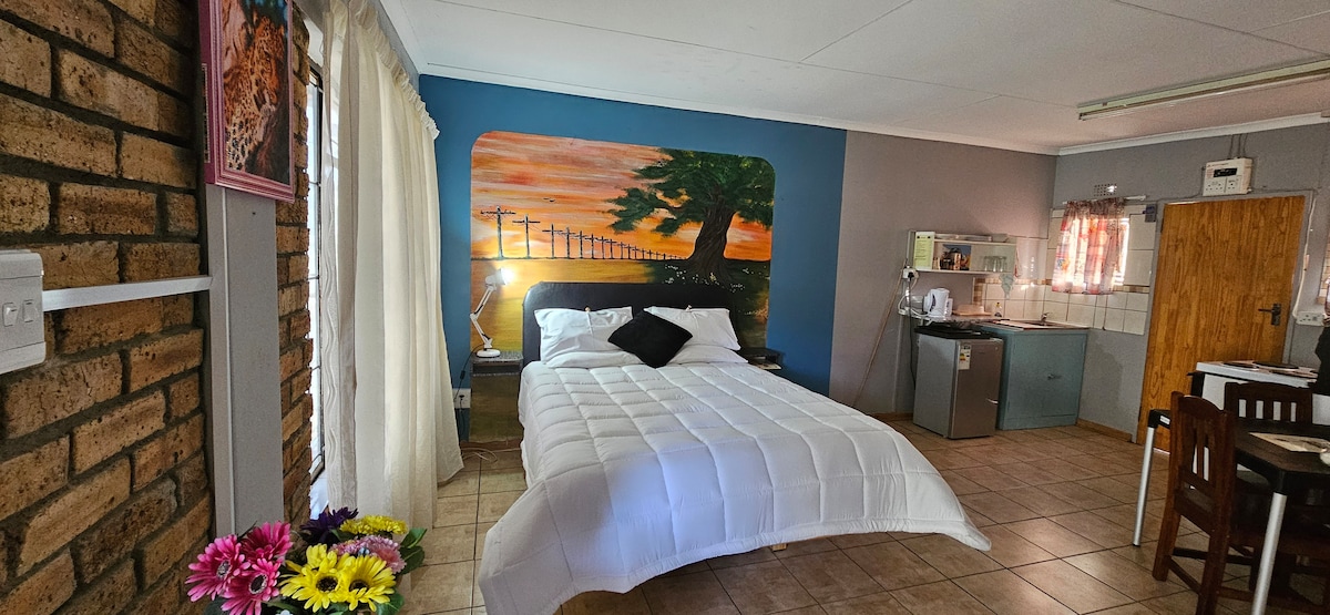 A spacious sleeping area is showcased with a large bed dressed in crisp white linen. A mural of a sunset and tree creates a striking backdrop. A kitchenette is visible to the side, with a dining table and wooden chairs nearby, complemented by colorful flowers.