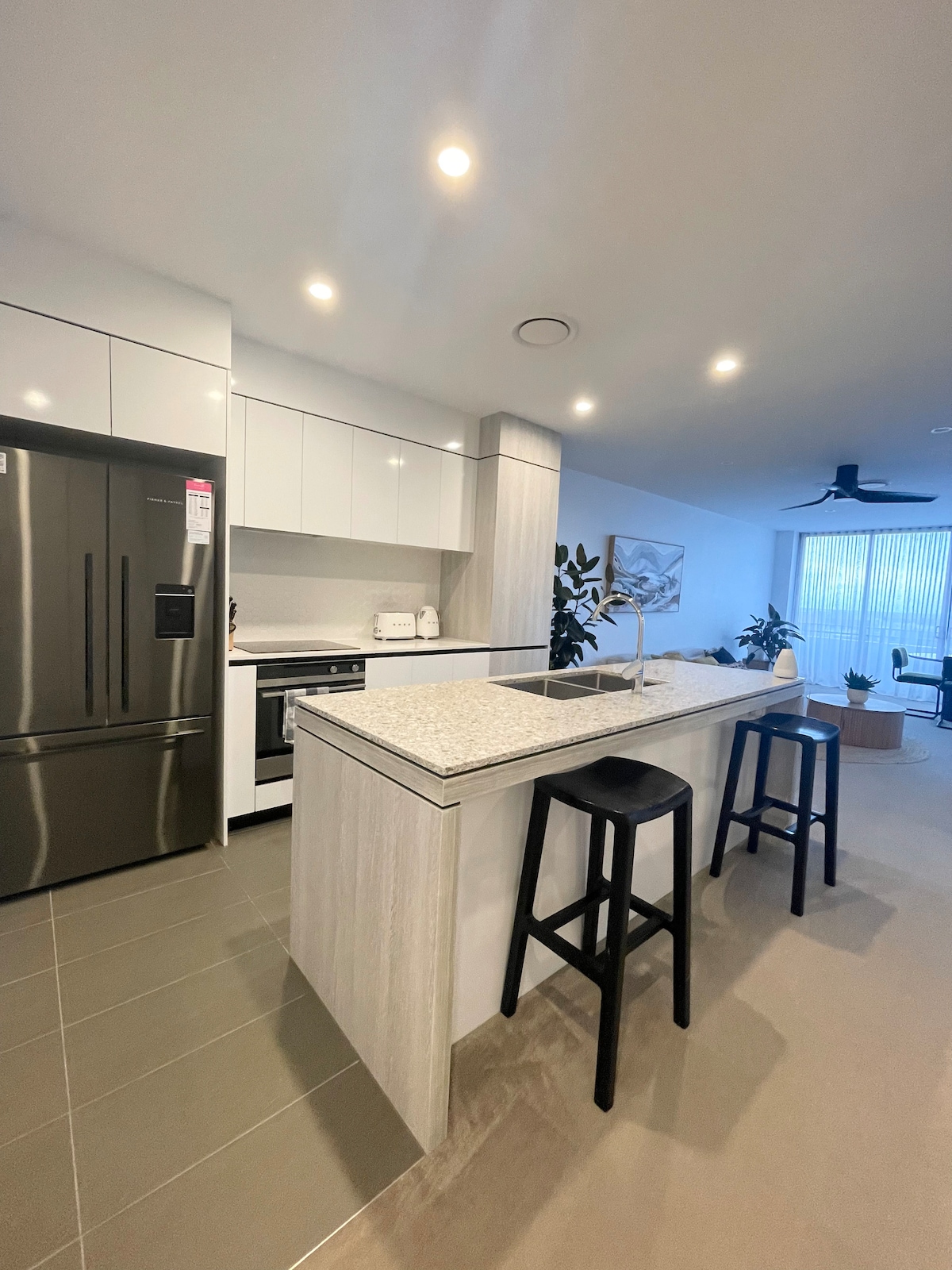 The modern kitchen is equipped with sleek, high-quality appliances, including a stainless-steel refrigerator and oven. A spacious island with a light stone countertop provides additional preparation space and seating with two black stools. Subtle cabinetry and potted plants enhance the overall ambiance.