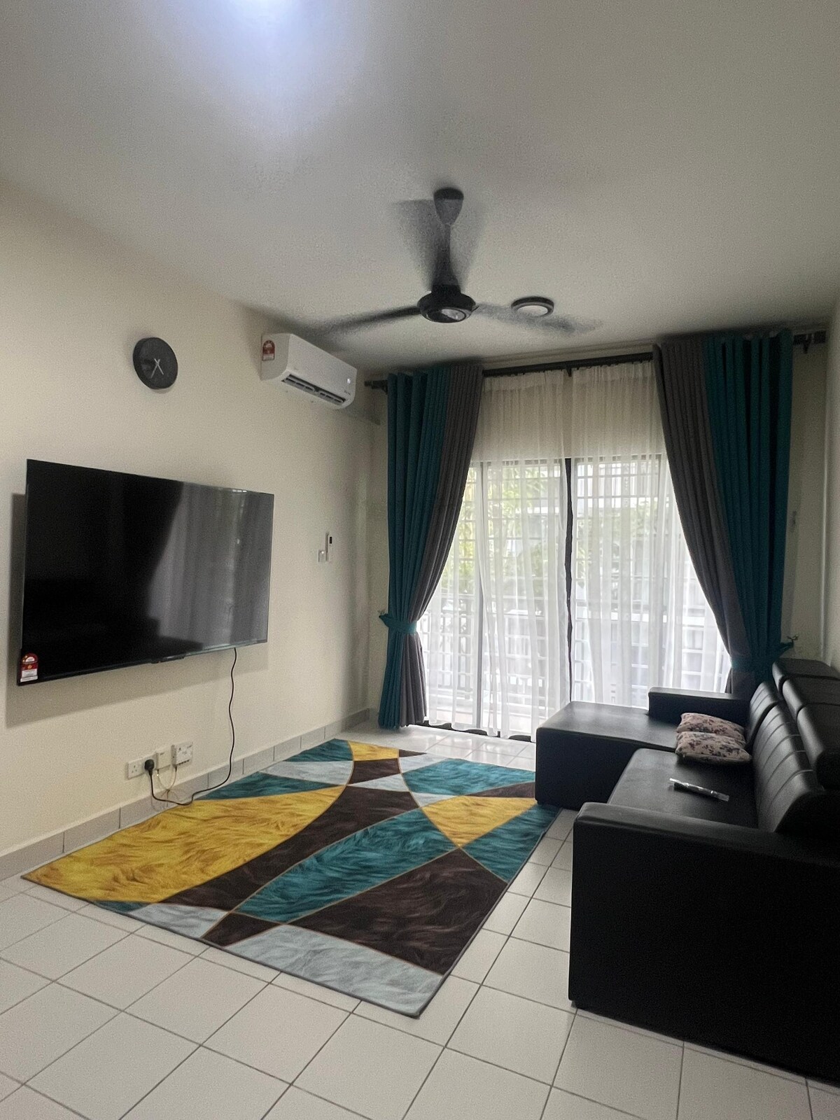 A living area features a black sectional sofa adjacent to a flat-screen television mounted on the wall. Light flows in through sheer curtains covering a sliding glass door, complemented by a colorful area rug that adds a vibrant touch to the tiled floor.