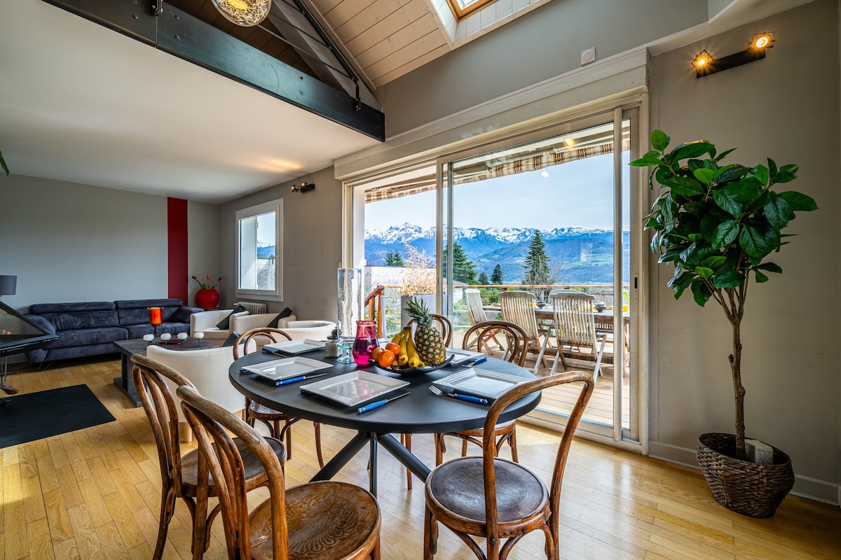 A dining area is visible with a large round table set for a meal, surrounded by wooden chairs. Large sliding doors open to a balcony, revealing a panoramic view of snow-capped mountains. Natural light illuminates the space, enhancing the modern décor.