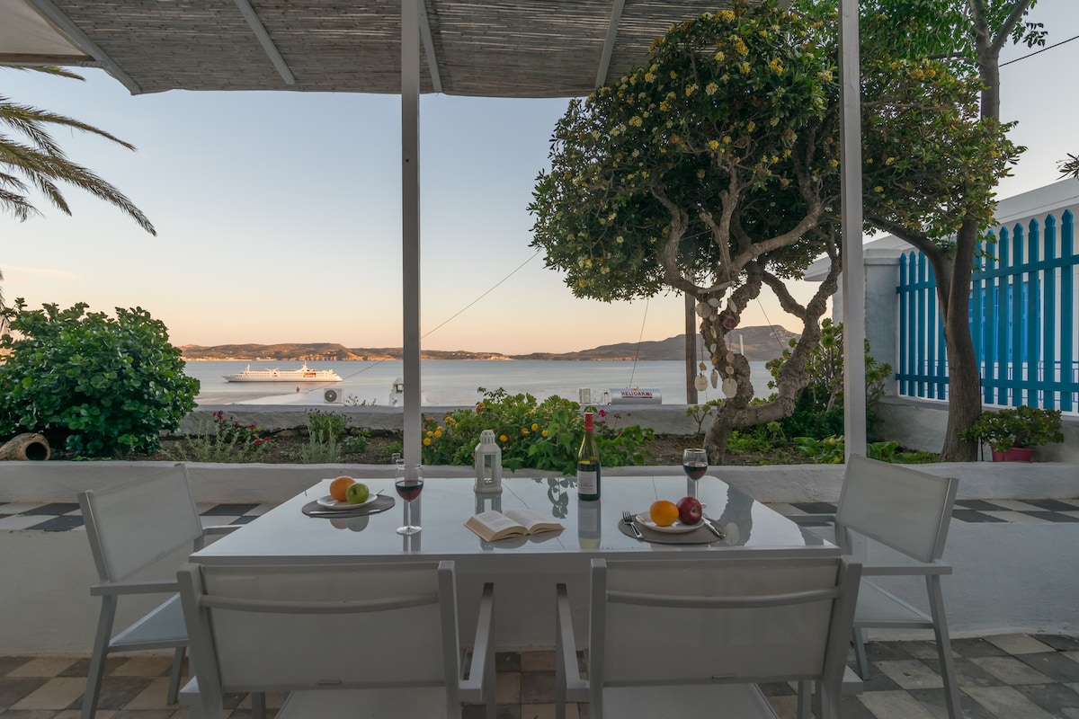 An outdoor terrace is equipped with a white dining table and four chairs, overlooking the Aegean Sea. A bottle of wine and glasses are visible, alongside fresh fruits. Lush greenery and palm trees frame the view, while colorful accents provide a vibrant backdrop.
