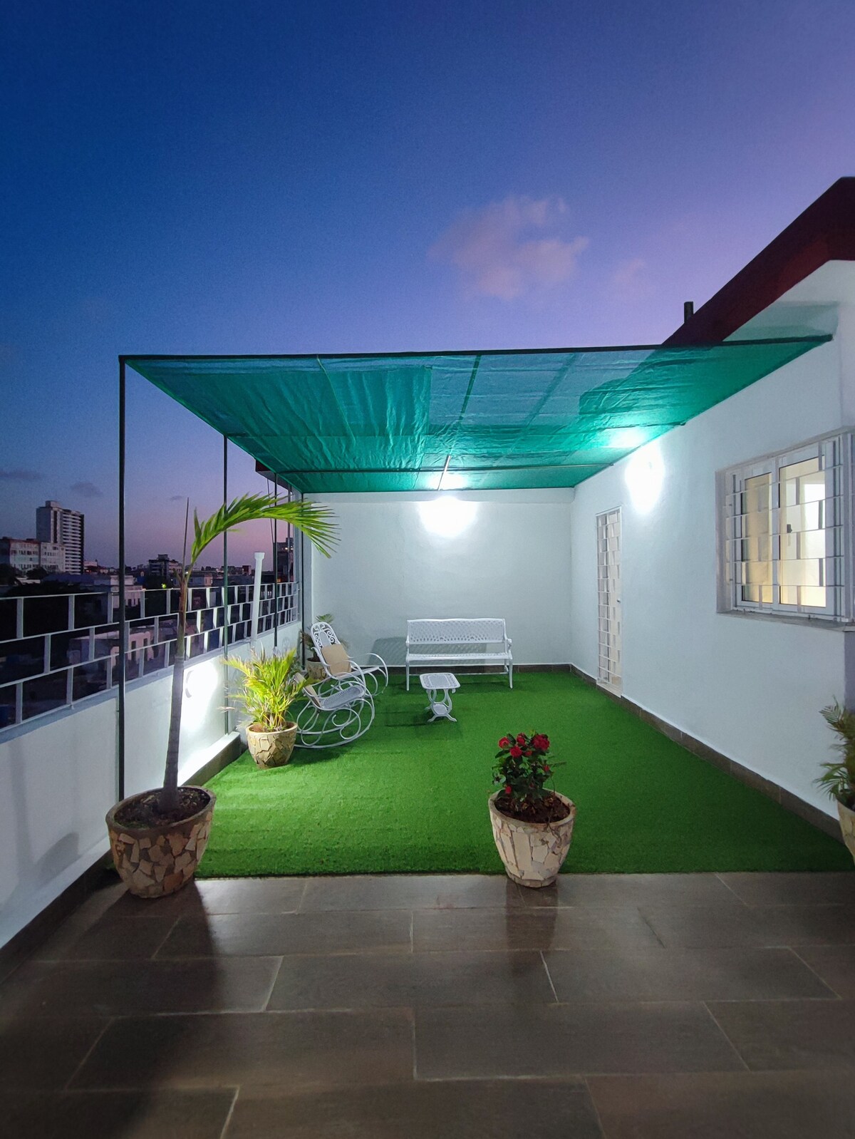 A private terrace features artificial grass underfoot, surrounded by potted plants. Two comfortable seating options are available, along with a side table. Soft lighting illuminates the space, enhancing the sense of tranquility as the evening sky transitions beyond.