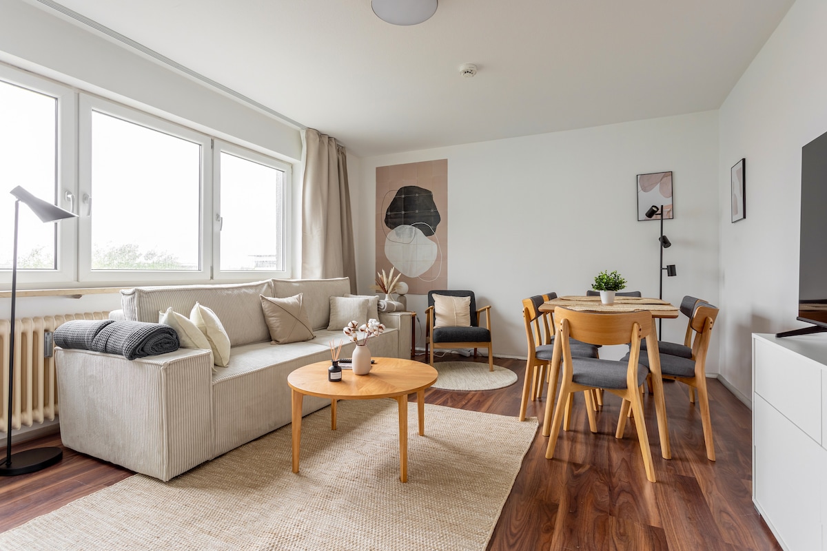 A modern living area features a light-colored sofa adorned with throw pillows. A round wooden coffee table sits centrally, surrounded by four dining chairs. A large rug anchors the space, with natural light streaming through two wide windows, illuminating the minimalist decor.