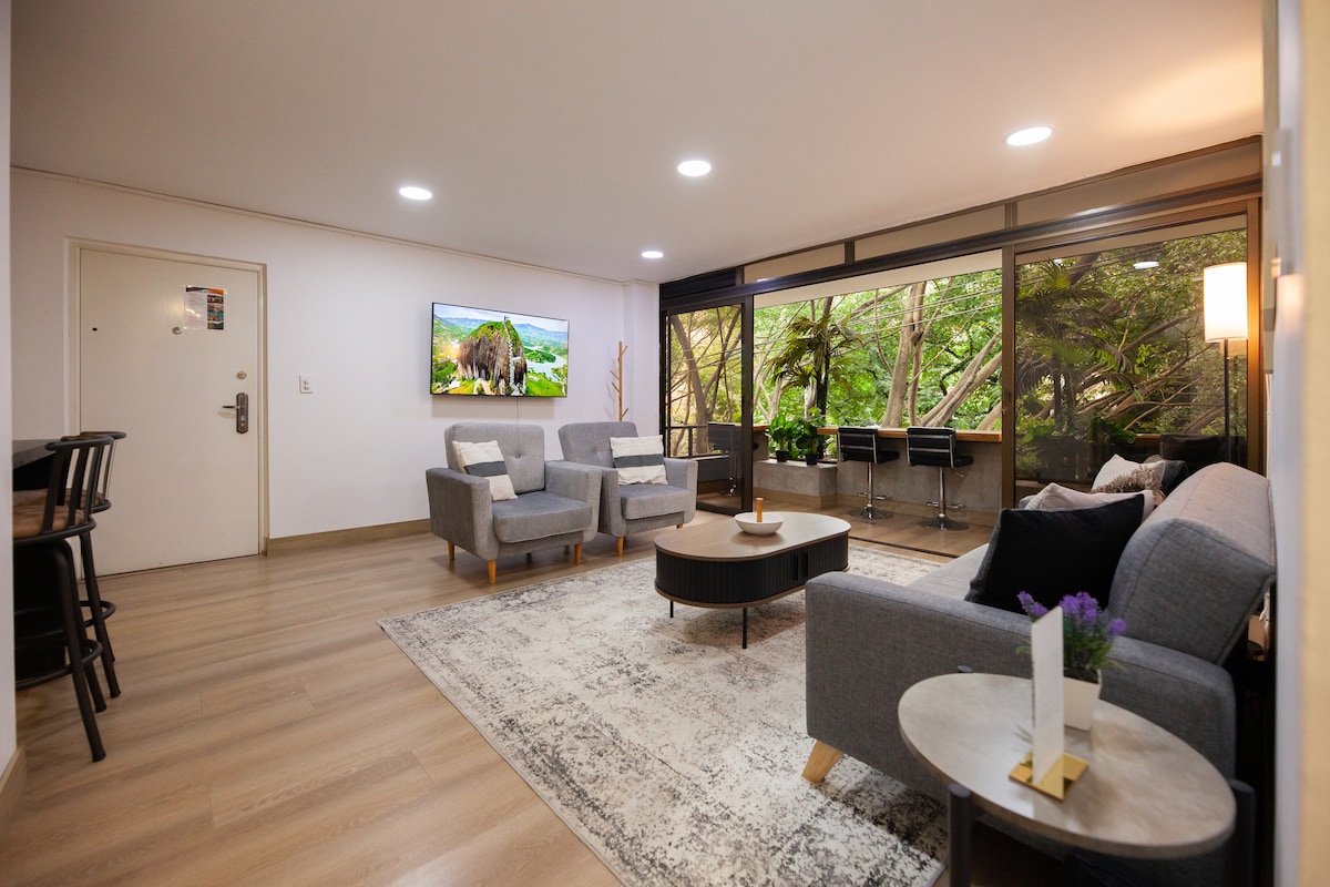 The spacious living room features modern furniture with a comfortable seating arrangement, including a sofa and armchairs. Large windows offer a view of lush greenery, while a stylish coffee table is positioned on a textured rug. A flat-screen TV is mounted on the wall.