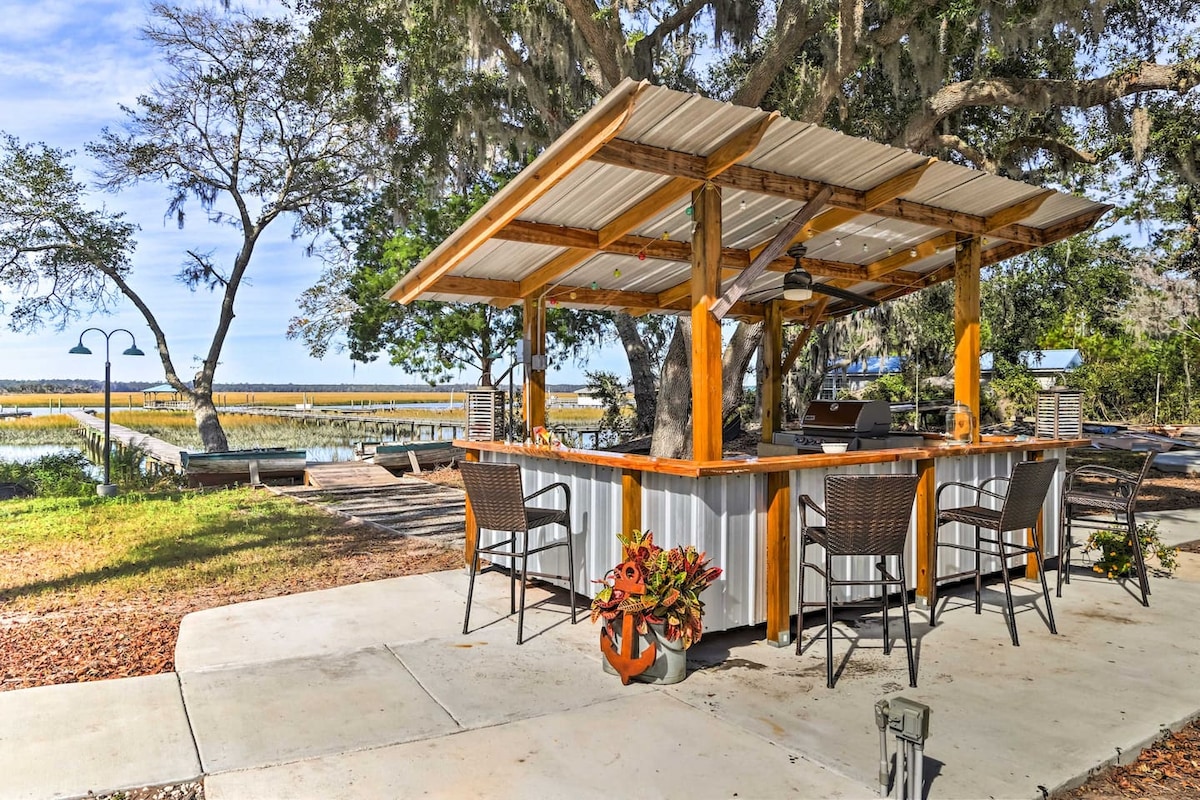 An outdoor kitchen area features a bar with several high-backed chairs. The rustic wooden structure is shaded with a metal roof, overlooking scenic water views. Potted plants add a touch of color, while a nearby dock enhances the serene atmosphere.