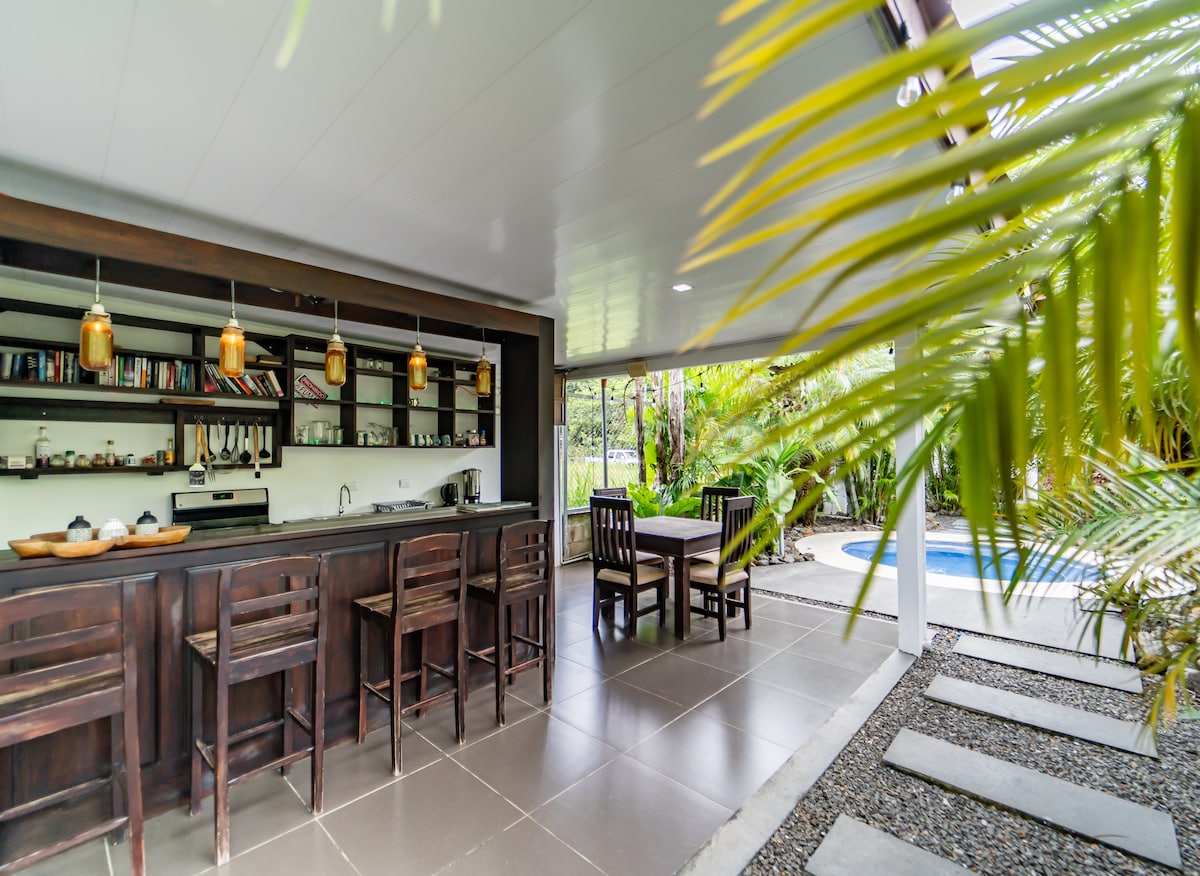 An outdoor bar area features dark wooden furniture, including high stools and a dining table surrounded by lush tropical plants. Shelves filled with various items are visible in the background, and a small, inviting jacuzzi is partially seen through the open space.