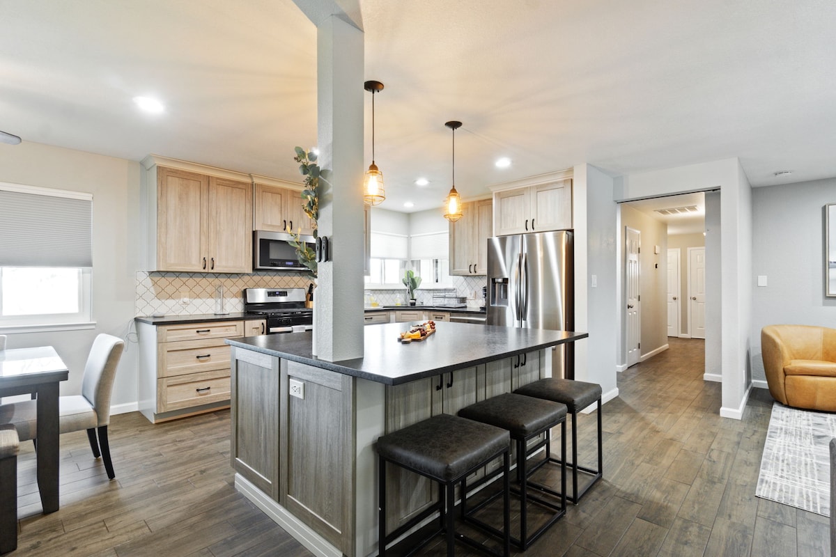 An open-concept kitchen showcases modern cabinetry and appliances, complemented by a large central island with seating for four. Pendant lights illuminate the space, while natural light streams through the windows, highlighting the wood floor and adjacent dining area.