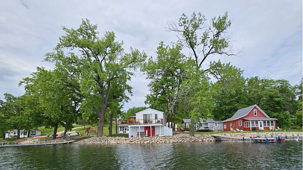 A tranquil lakeside scene showcases two charming cabins nestled among lush green trees. The elevated cabin features a spacious balcony overlooking the water, while the neighboring cabin displays a welcoming red facade. A pebble beach leads to a dock, inviting leisure activities.
