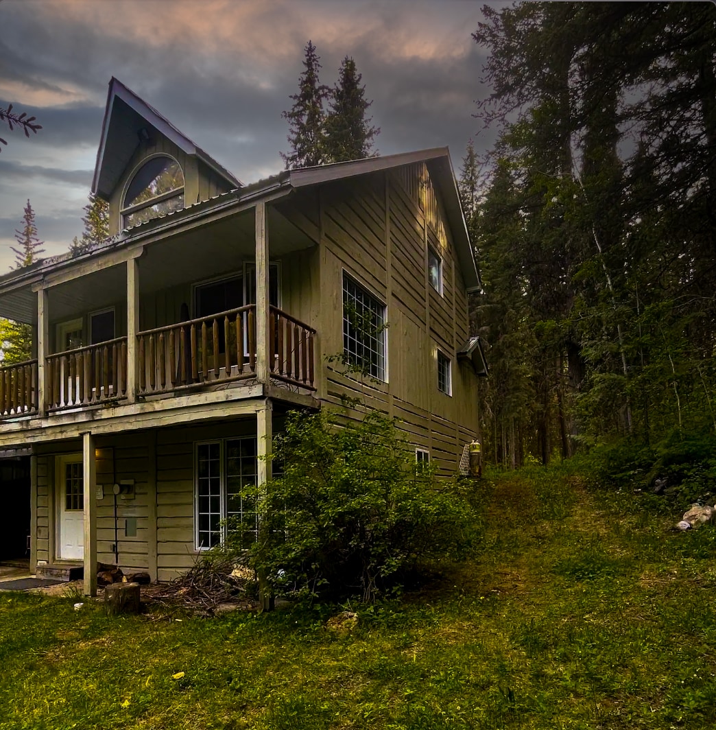 The cabin's exterior showcases a two-story structure with a sloped roof and a balcony, surrounded by lush greenery. Tall trees flank the building, and a pathway leads through the grassy area, inviting guests to explore the natural surroundings.