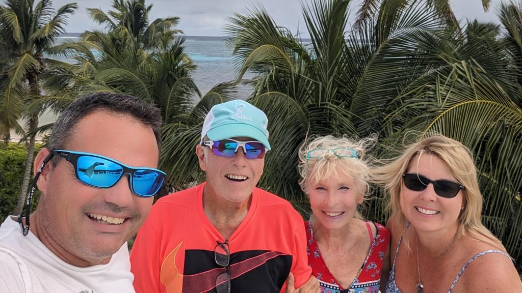 A group of four individuals is captured in front of lush palm trees, with the Caribbean Sea visible in the background. The scene conveys a relaxed atmosphere, enhanced by the gentle sea breeze, indicative of a beachfront retreat.
