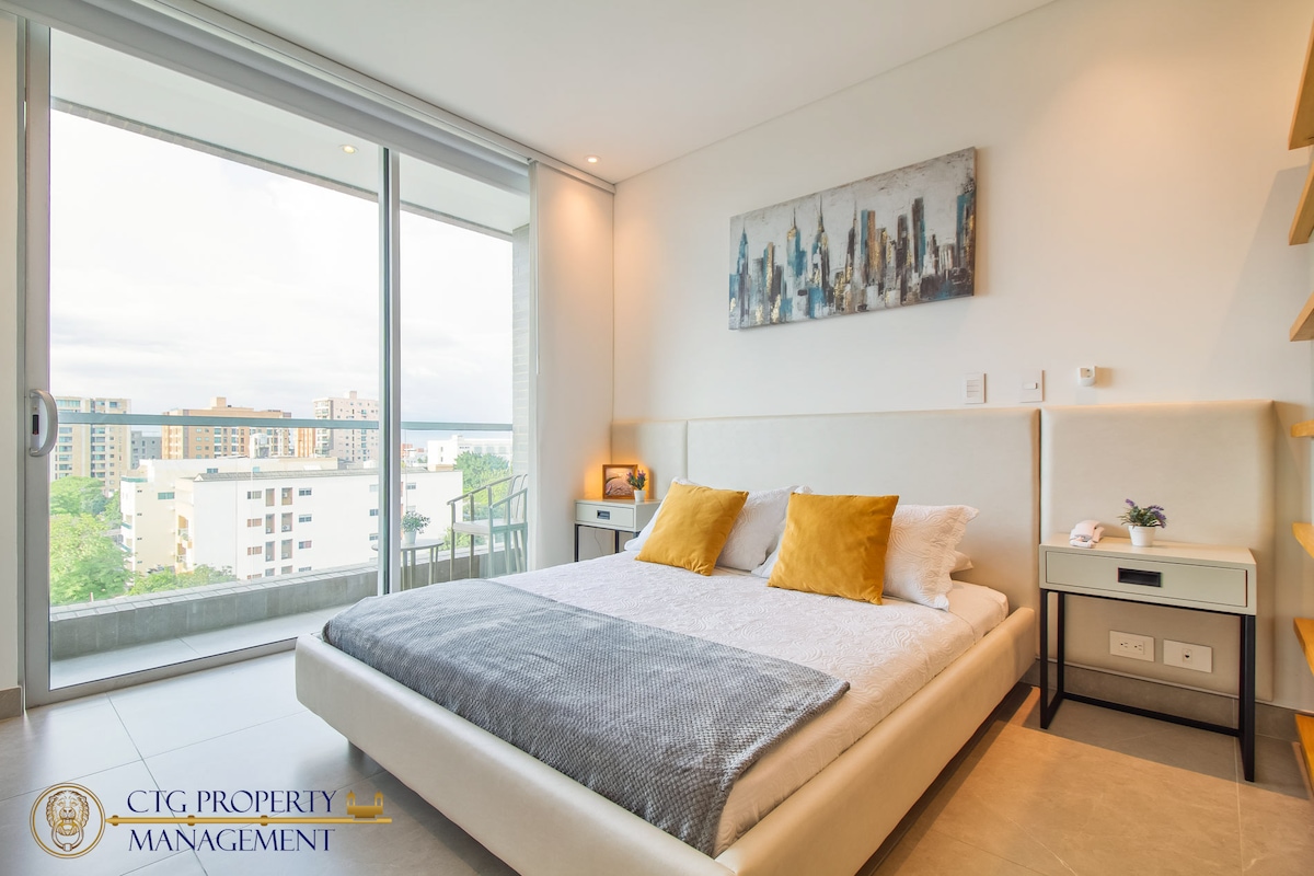 A spacious bedroom features large glass doors leading to a balcony, providing natural light and a view of the city. The bed is dressed in soft linens with decorative pillows. Subtle shelving units are positioned beside a bedside table, enhancing the room's functionality.