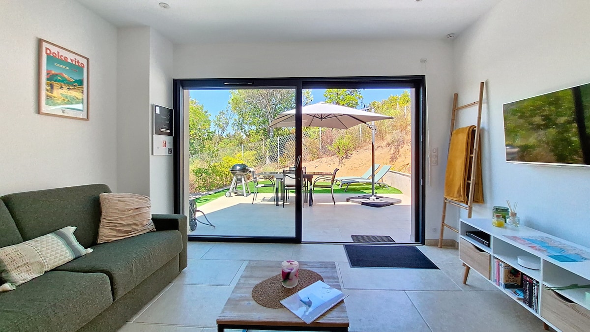 A living area features a spacious green sofa facing sliding glass doors leading to a private terrace. A coffee table sits in front, while a flat-screen TV is mounted on the wall. The patio outside includes outdoor seating and a parasol, surrounded by greenery.