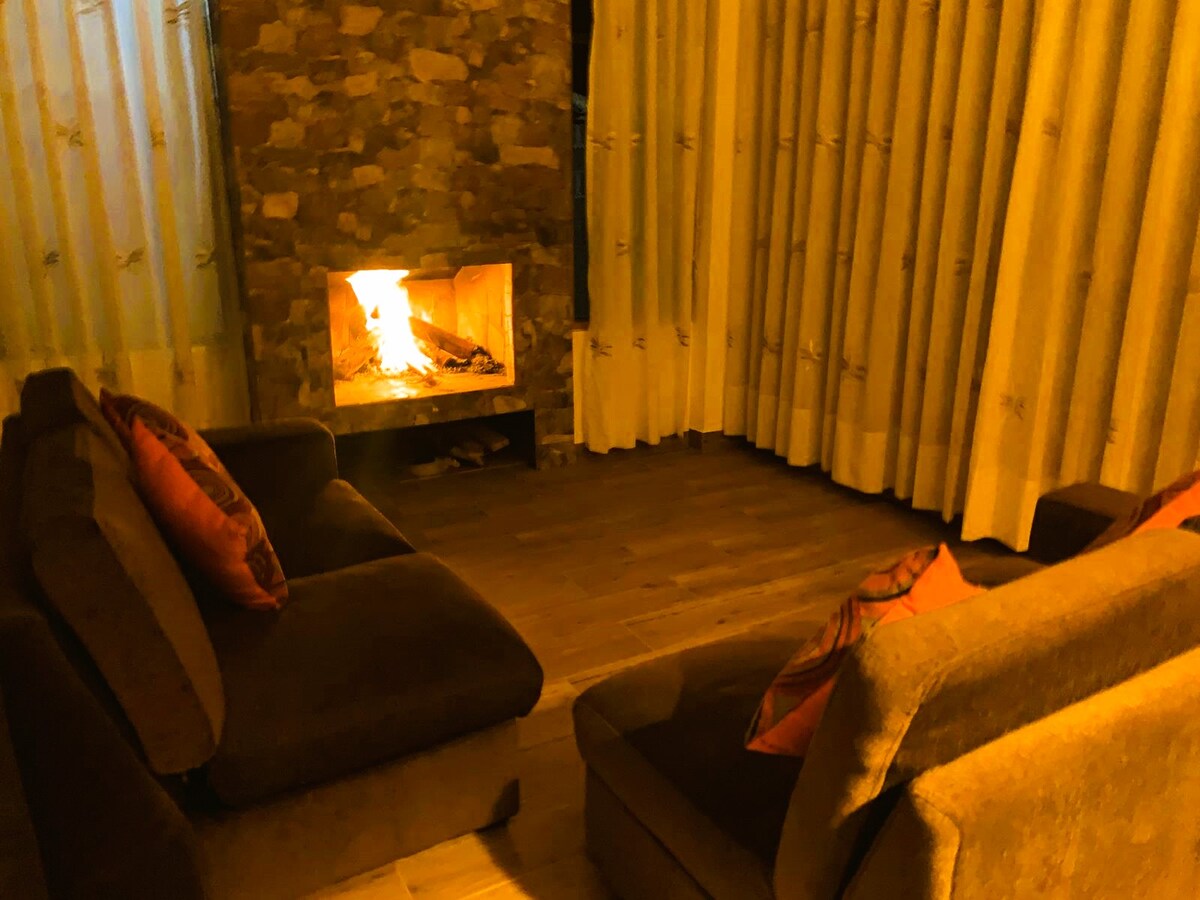 A warm living area features two comfortable sofas with accent pillows, positioned near a stone fireplace. Soft lighting from the fire creates a cozy atmosphere, while long curtains frame the scene, enhancing the sense of comfort and relaxation.