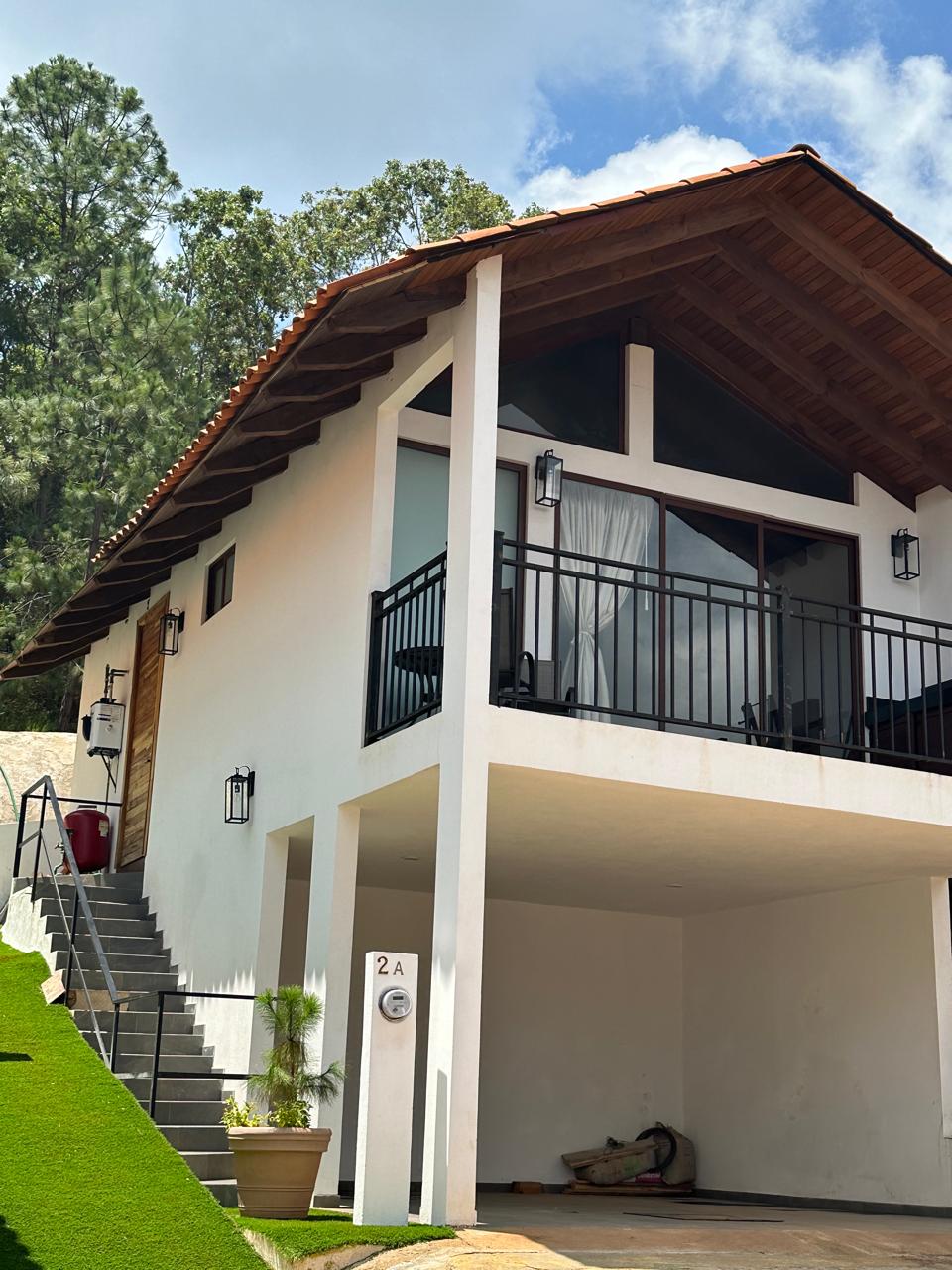 The exterior of a two-story structure features a sloped roof and a large balcony, with two sets of steps leading to the entrance. A small potted plant is positioned at the base of the steps, and the grassy area enhances the surrounding landscape.