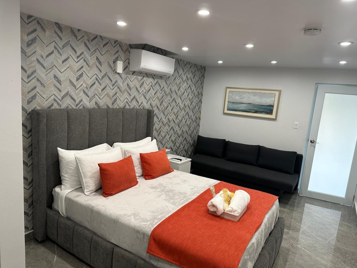 A spacious bedroom showcases a large upholstered bed adorned with white linens and orange accent pillows. A modern sofa is positioned against the wall, with a decorative artwork above the bed. Soft lighting enhances the clean, contemporary design.