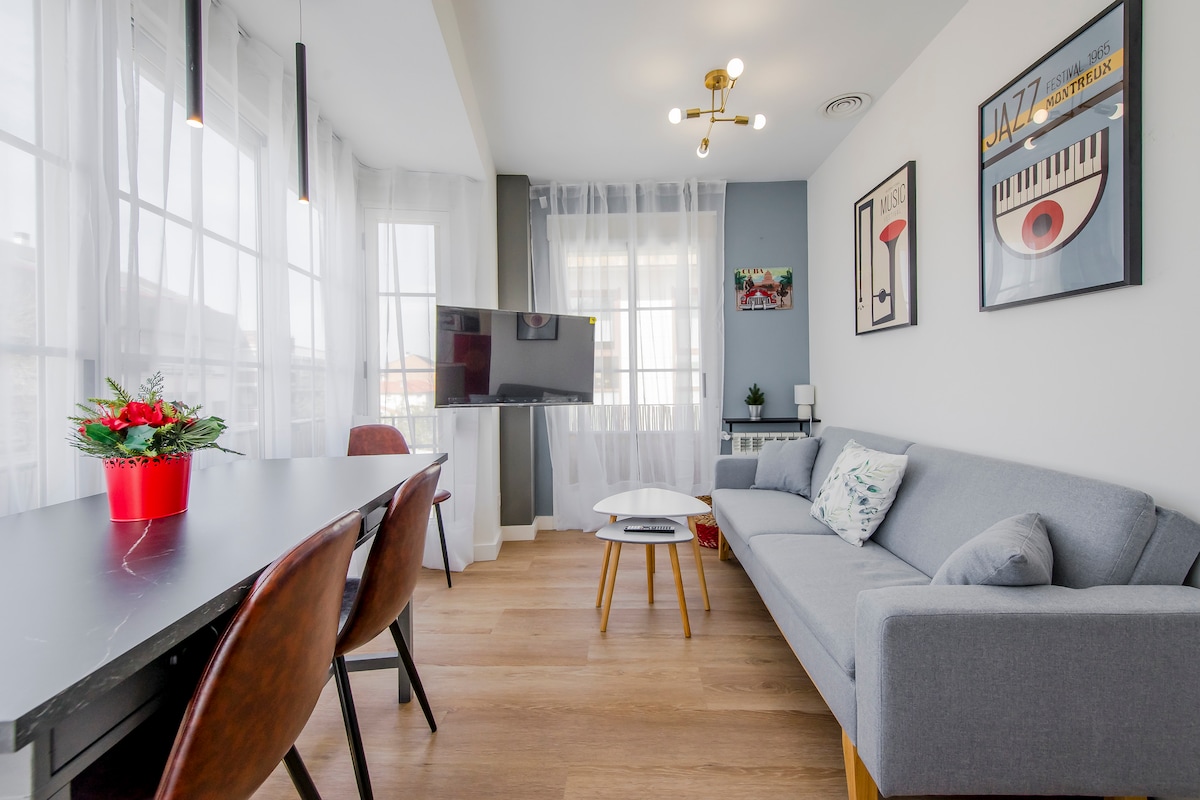A modern open-plan living space is featured, showcasing a comfortable gray sofa and a sleek white coffee table. Large windows allow natural light to fill the room, while decorative plants and framed art pieces add a touch of charm. A flat-screen TV is mounted on the wall.