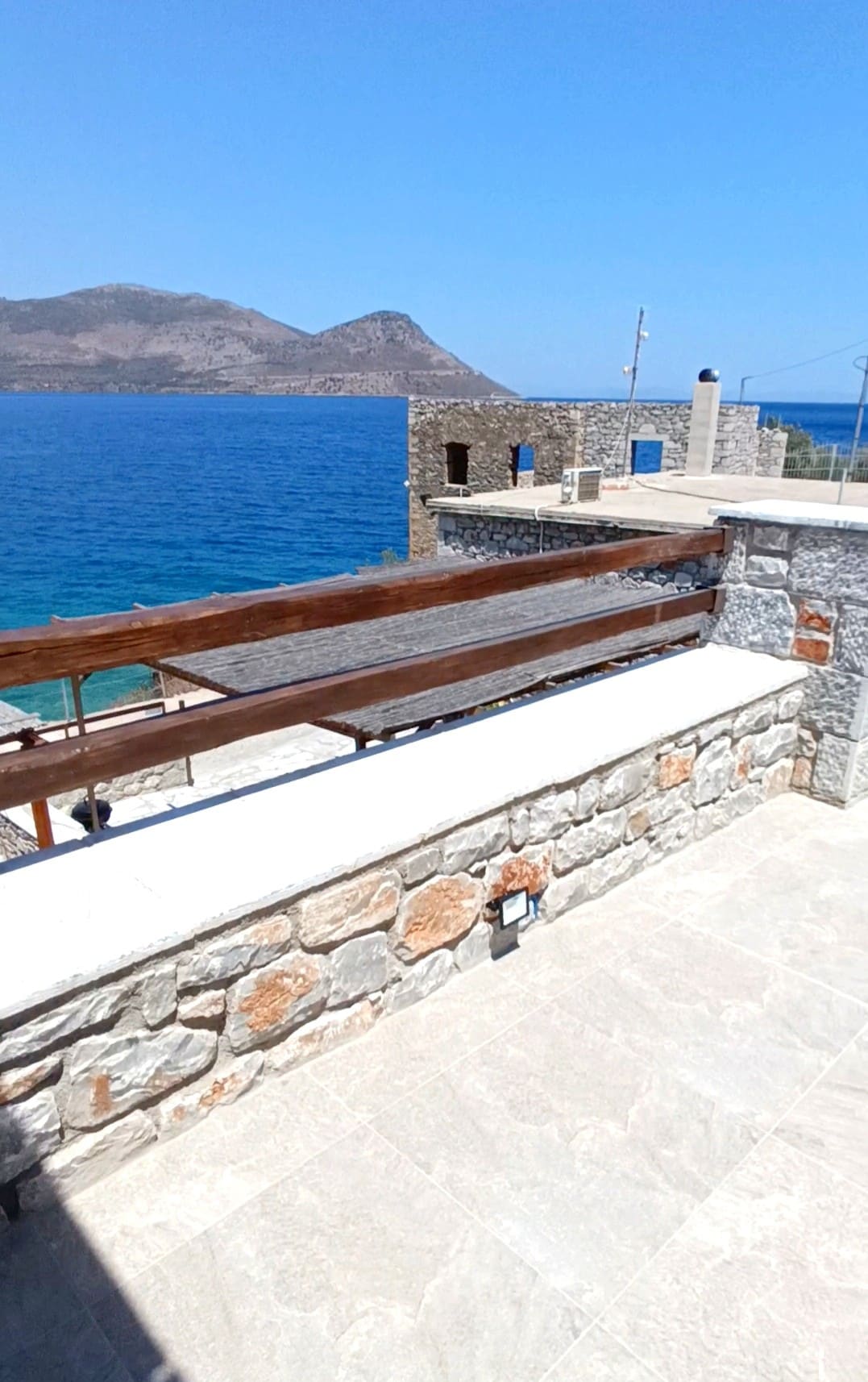 A spacious terrace features natural stone walls and wooden railings, overlooking the sea. The serene blue waters reflect the clear sky, while a distant mountain enhances the scenic view. The smooth stone surface provides a comfortable area for relaxation.