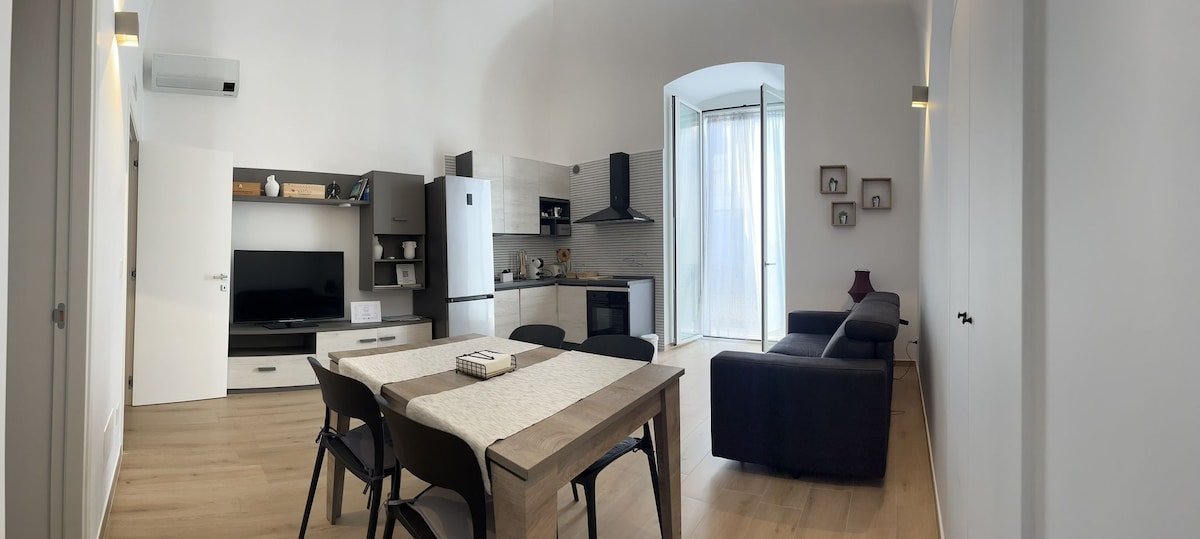 An open living area showcases a contemporary design with large windows that allow natural light. A dining table is accompanied by black chairs, while a dark sofa is positioned nearby. The kitchen area features modern appliances, including a stovetop and a television.