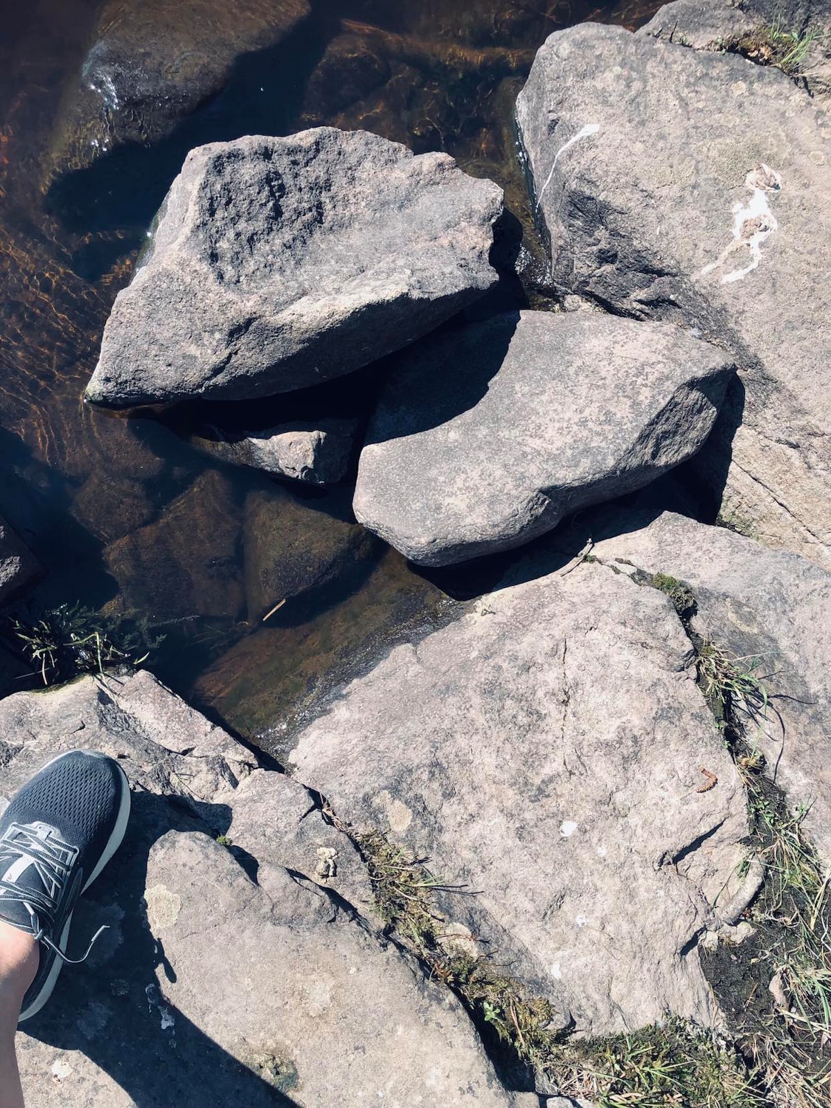 Large, flat stones are partially submerged in clear water, creating a natural stepping path. Green grass is visible on the edges, contrasting with the rough texture of the rocks. A person’s foot is shown on one stone, suggesting the tranquility of the surrounding water.