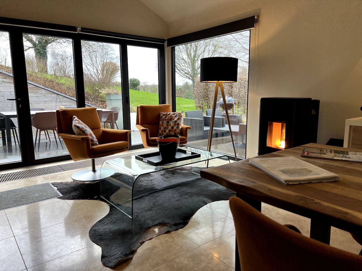 A spacious living area features large glass doors that invite natural light and views of the outdoors. Two cozy armchairs are positioned around a sleek glass coffee table, with a soft area rug beneath. A modern floor lamp and a pellet stove add comfort to the space.