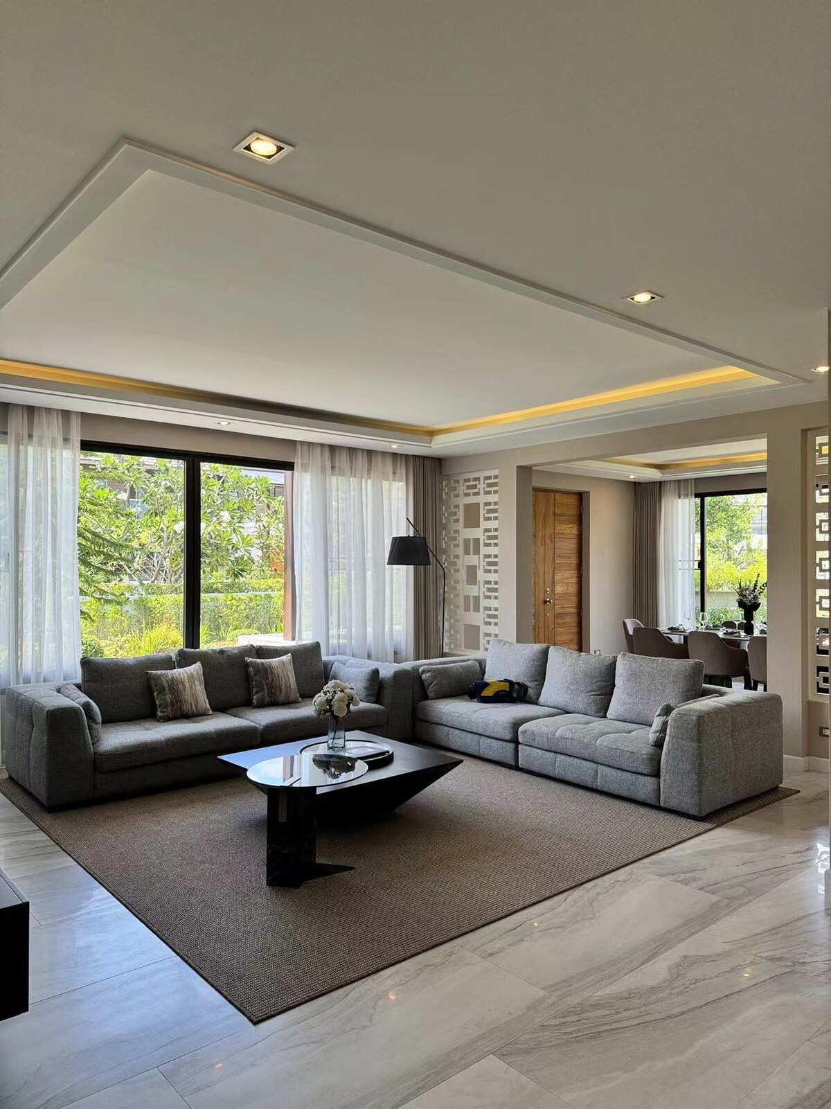 A spacious living area features two comfortable sofas arranged around a sleek coffee table, set on a large area rug. Large windows allow natural light to illuminate the room and offer views of the greenery outside.