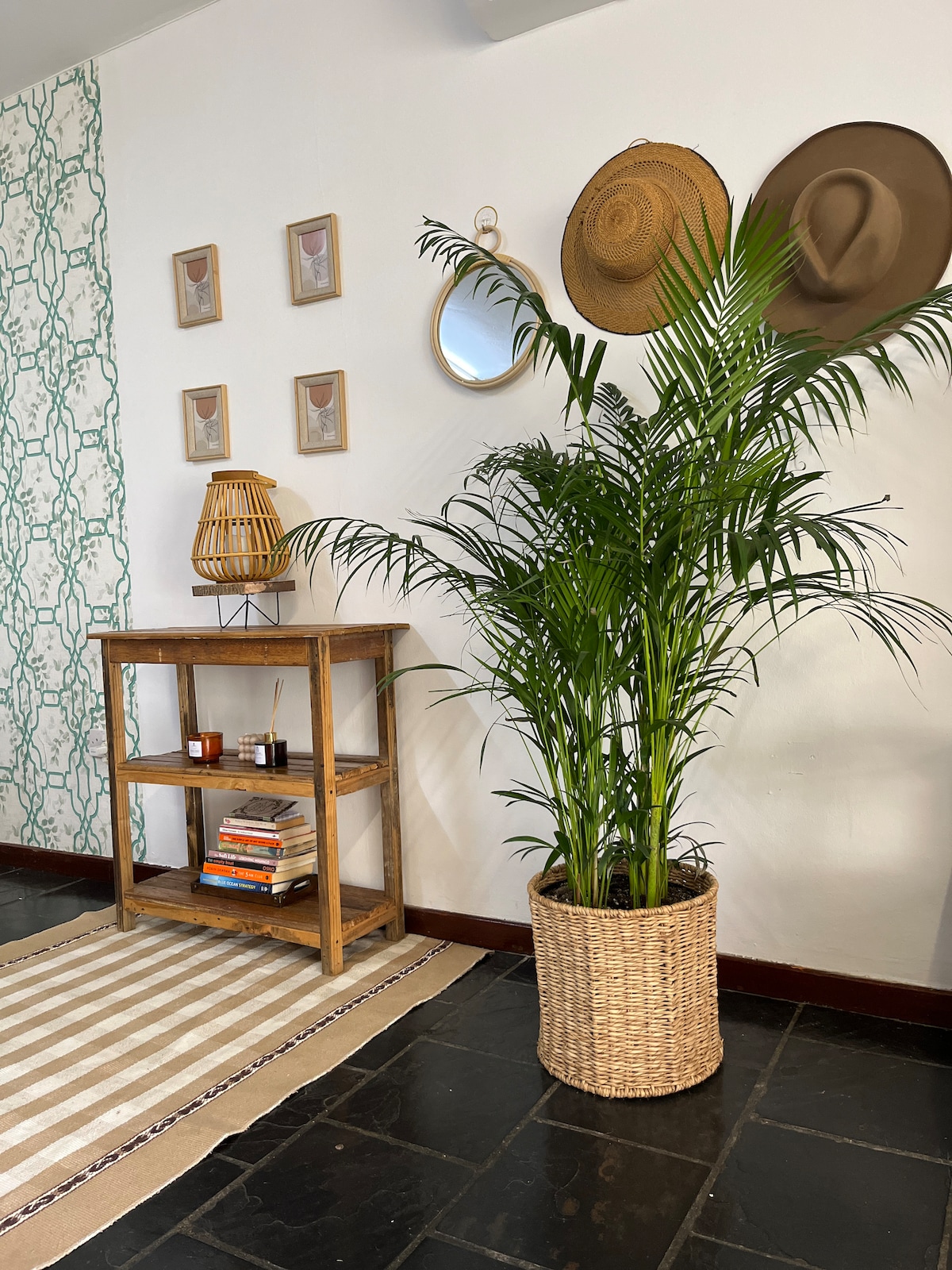 A decorative corner features a wooden shelving unit holding books and candles. A tall, leafy plant is presented in a woven basket, complementing the space. Framed pictures and hats hang on the wall, which is adorned with a subtle patterned design.
