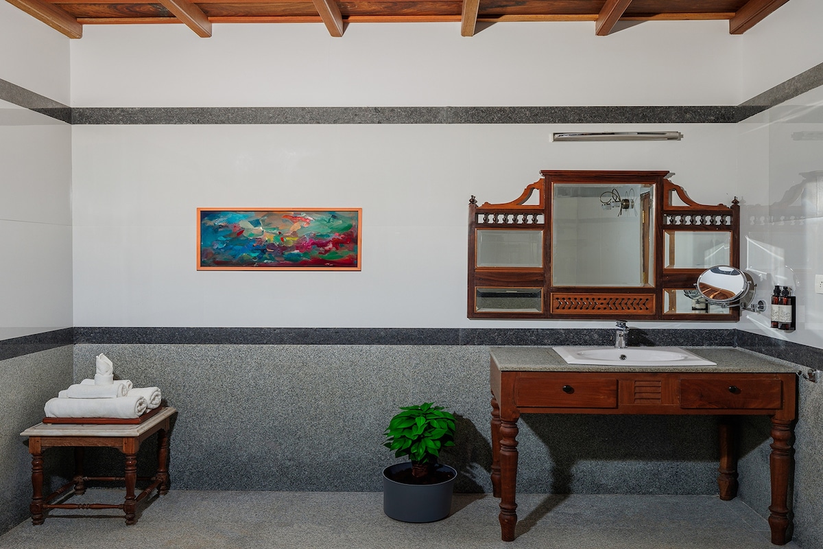 A spacious bathroom is highlighted by a wooden vanity with a sink and a mirror above. Fresh towels are neatly arranged on a stool. A vibrant artwork adds color to the wall, while a potted plant introduces a touch of nature.