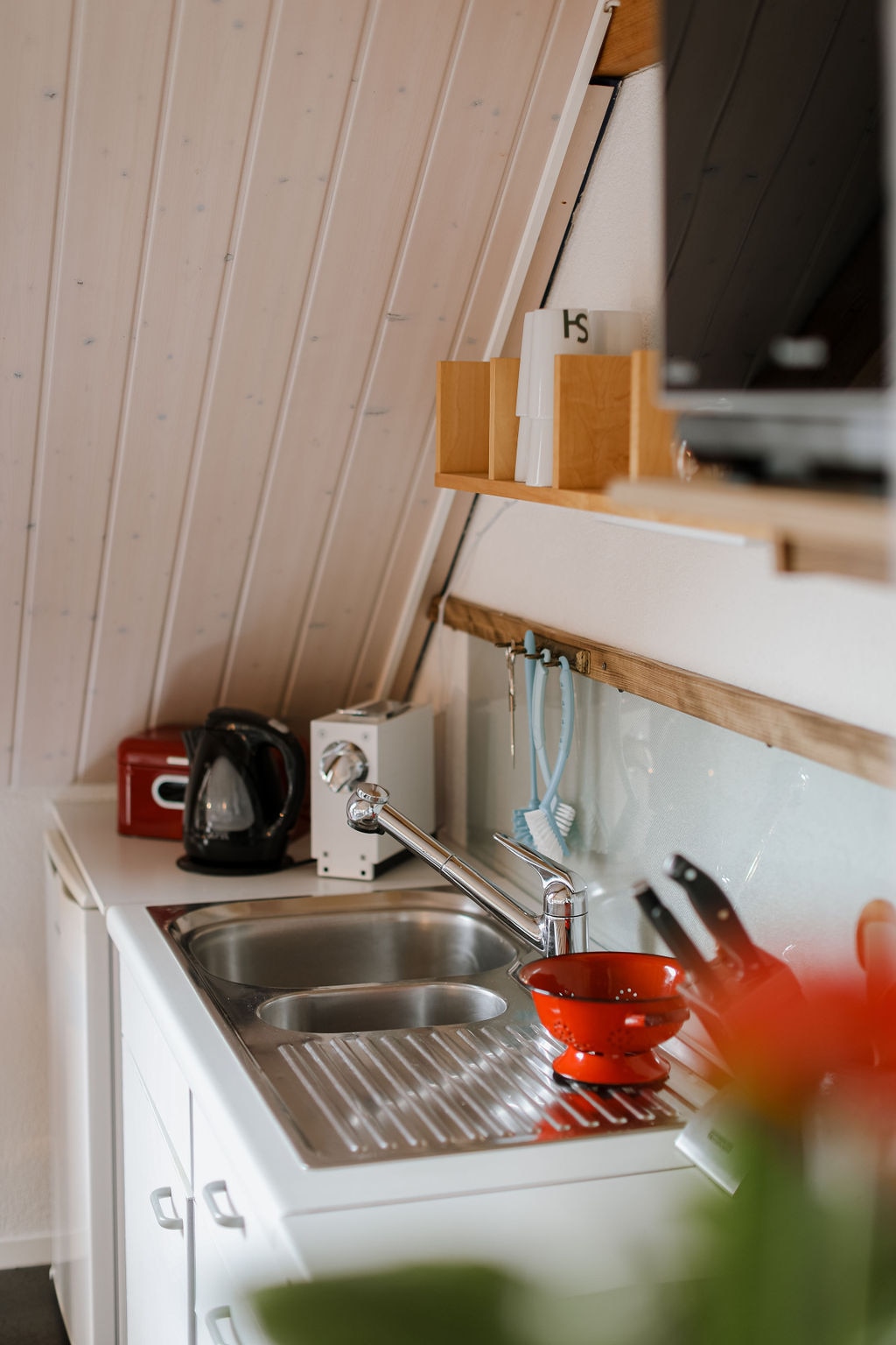 A compact kitchen area features a metal sink with a draining board, alongside a red kettle and a white toaster. Kitchen utensils are arranged neatly, with a small red bowl placed prominently on the countertop. Natural light reflects off the white cabinetry and sloped ceiling.