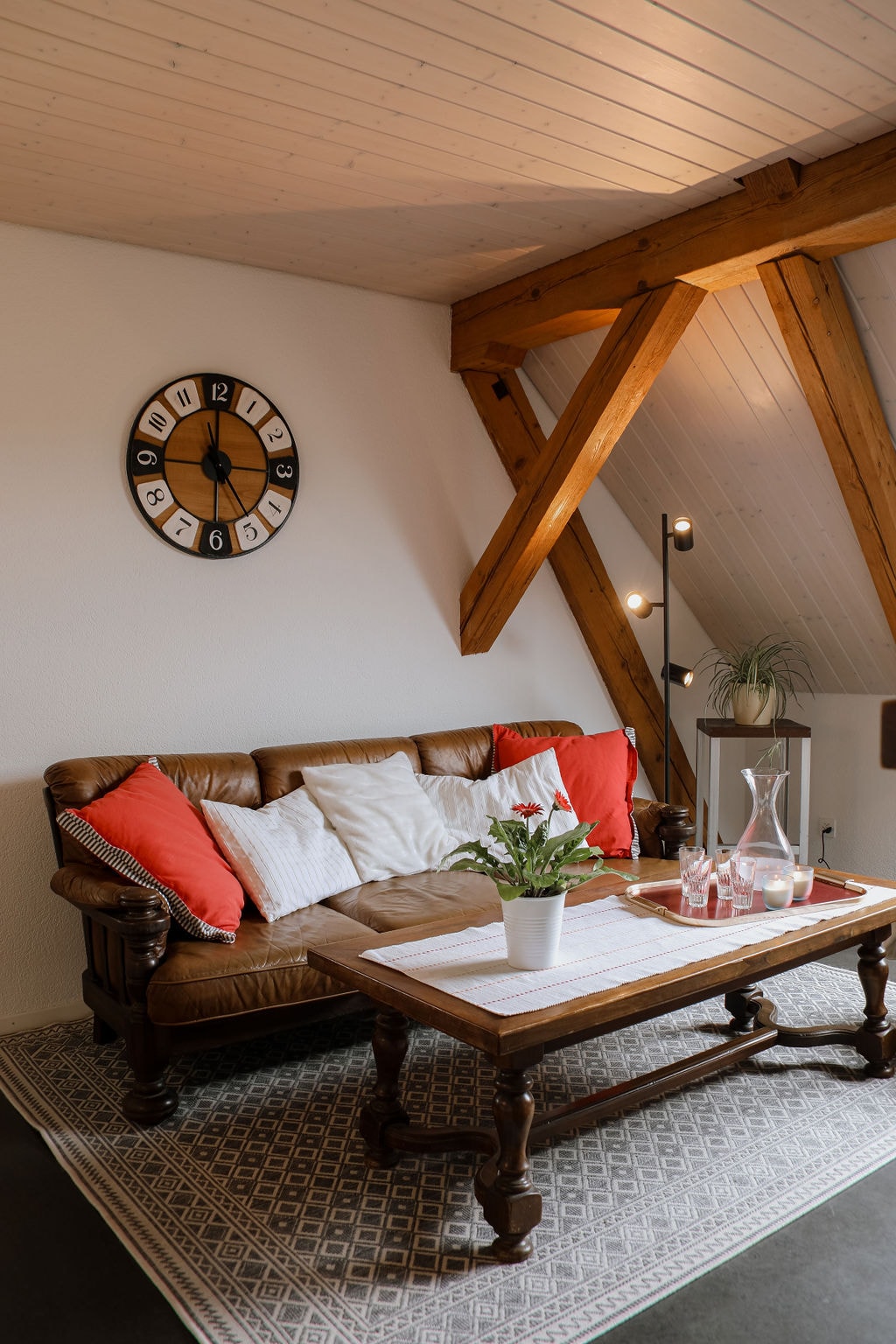 A cozy seating area features a brown sofa adorned with red cushions. A wooden table is set in front, displaying a potted plant and glassware. Light wood beams create an airy atmosphere, with a clock mounted on the wall, adding a functional element to the space.