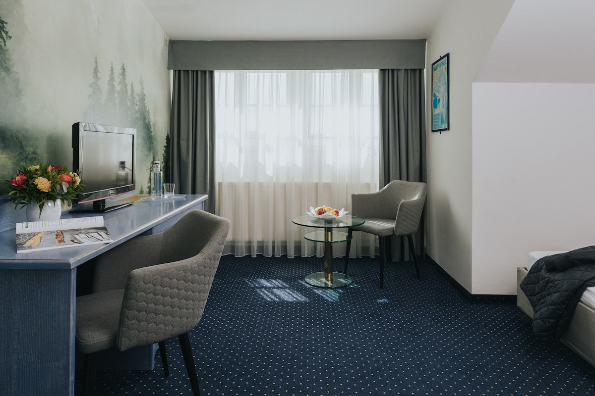 A modern room features a small table with fresh fruit and two comfortable chairs. A desk is positioned nearby, with a flat-screen TV mounted on the wall. Light filters through sheer curtains, softly illuminating the space, while a nature-themed mural adds a serene touch.