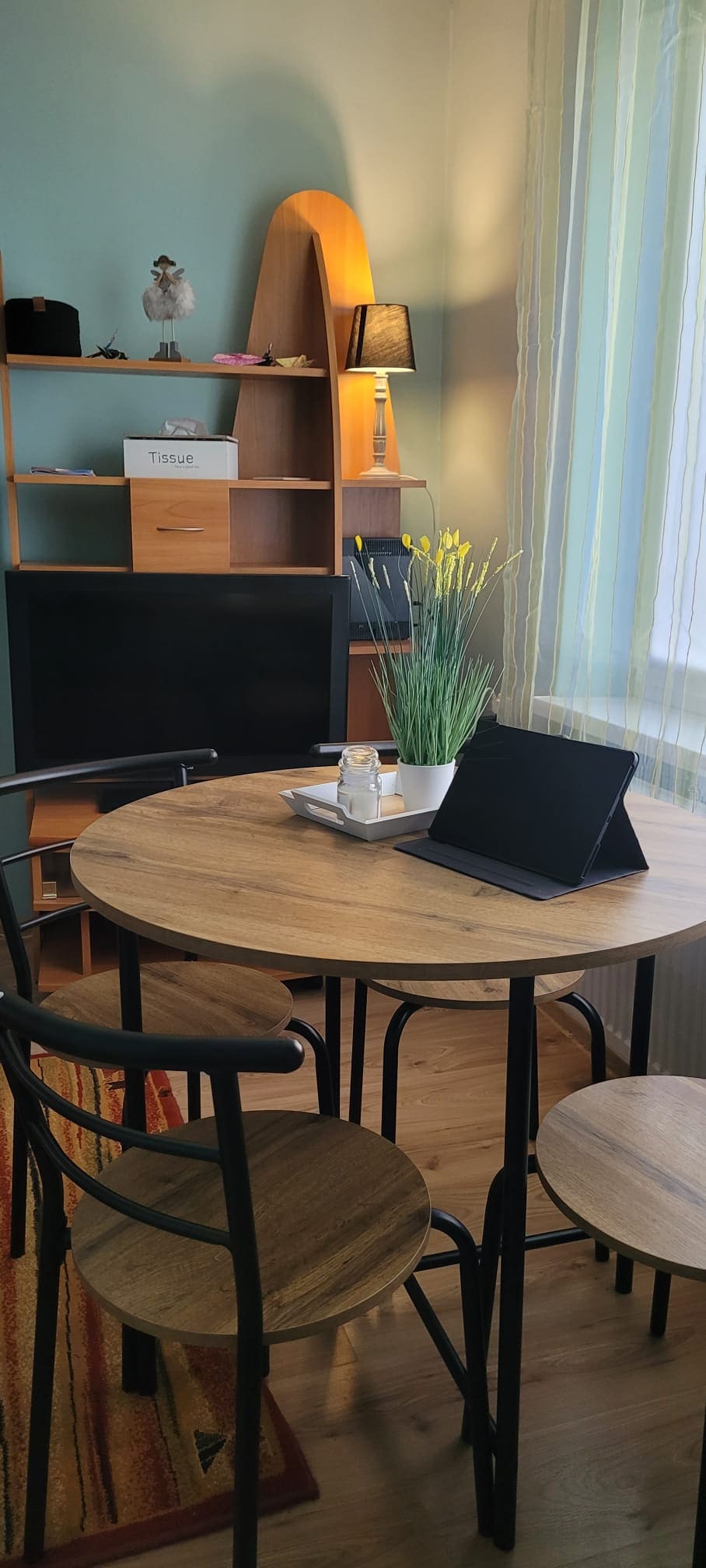 A round wooden dining table is surrounded by four black chairs, two with backs and two without. A tablet stands on the table beside a small vase filled with yellow flowers. Natural light enters through sheer curtains, illuminating a shelving unit in the background.