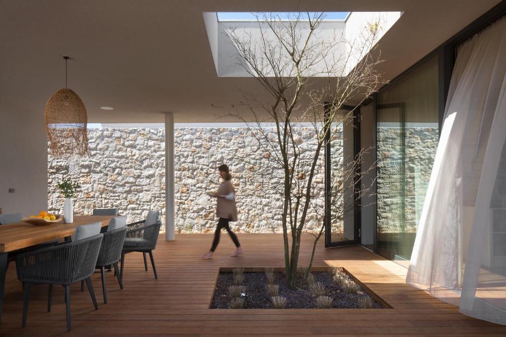 A modern outdoor living area features a central decorative tree surrounded by small stones. A wooden dining table with six gray wicker chairs is positioned nearby. Large sliding glass doors and a skylight allow natural light to flood the space, enhancing the tranquil atmosphere.
