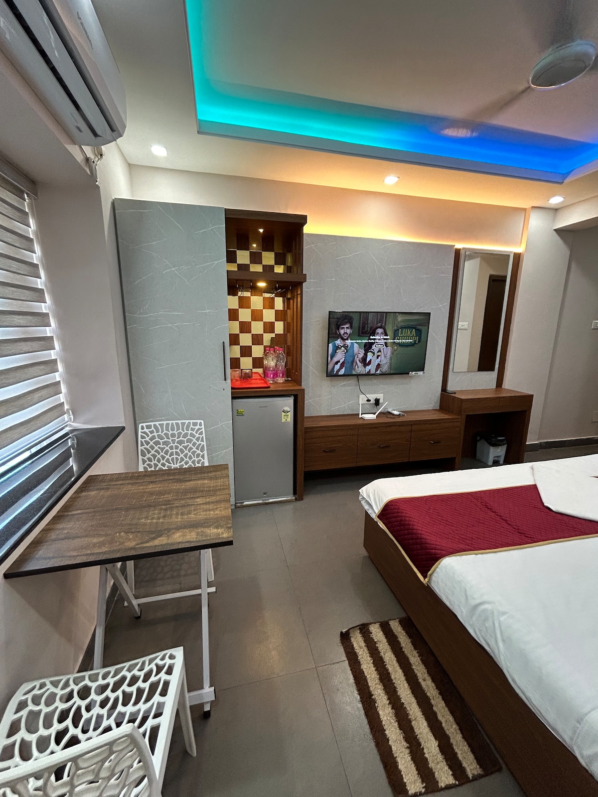 A well-appointed room features a spacious bed with a contrasting red throw. A small kitchenette with modern appliances is visible, along with a TV mounted on the wall. A sleek wooden table and two chairs provide dining space, enhanced by soft, ambient lighting.