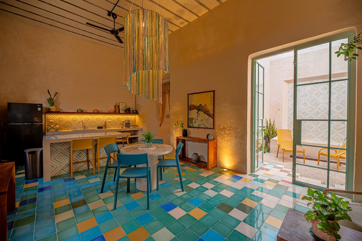 An open dining area features a round table surrounded by colorful chairs. Unique tiled flooring showcases a blend of blue and green. A large window allows natural light to illuminate the space, while decorative plants add a touch of greenery.