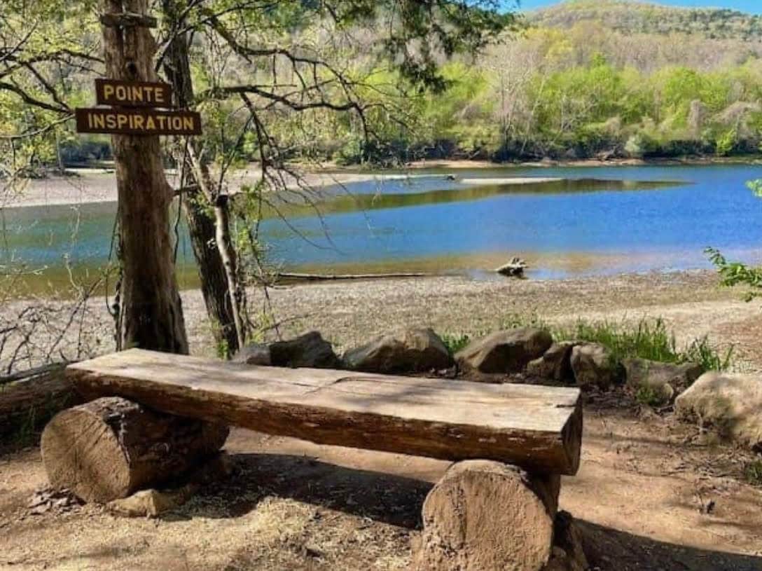 A rustic wooden bench is positioned beside a calm water body, surrounded by greenery. A sign labeled 'Pointe Inspiration' is mounted on a nearby tree. The scene captures a tranquil setting, with hills visible in the background and gentle ripples on the water's surface.