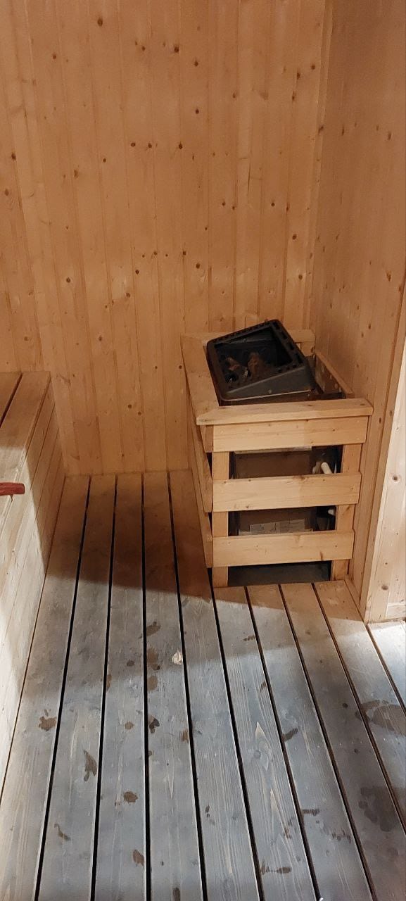 A wooden sauna interior features light-toned wooden paneling and floorboards. A small wooden bench is positioned alongside a wooden storage unit, which holds a black heating element. The atmosphere of warmth and relaxation is suggested by the sauna’s design.