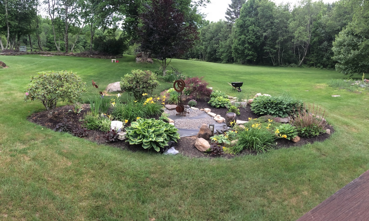 A vibrant garden is featured, surrounded by lush green grass and mature trees. The circular flower bed showcases a variety of plants, including colorful flowers and ornamental stones, creating a serene outdoor space for relaxation. A fire pit is visible in the background.