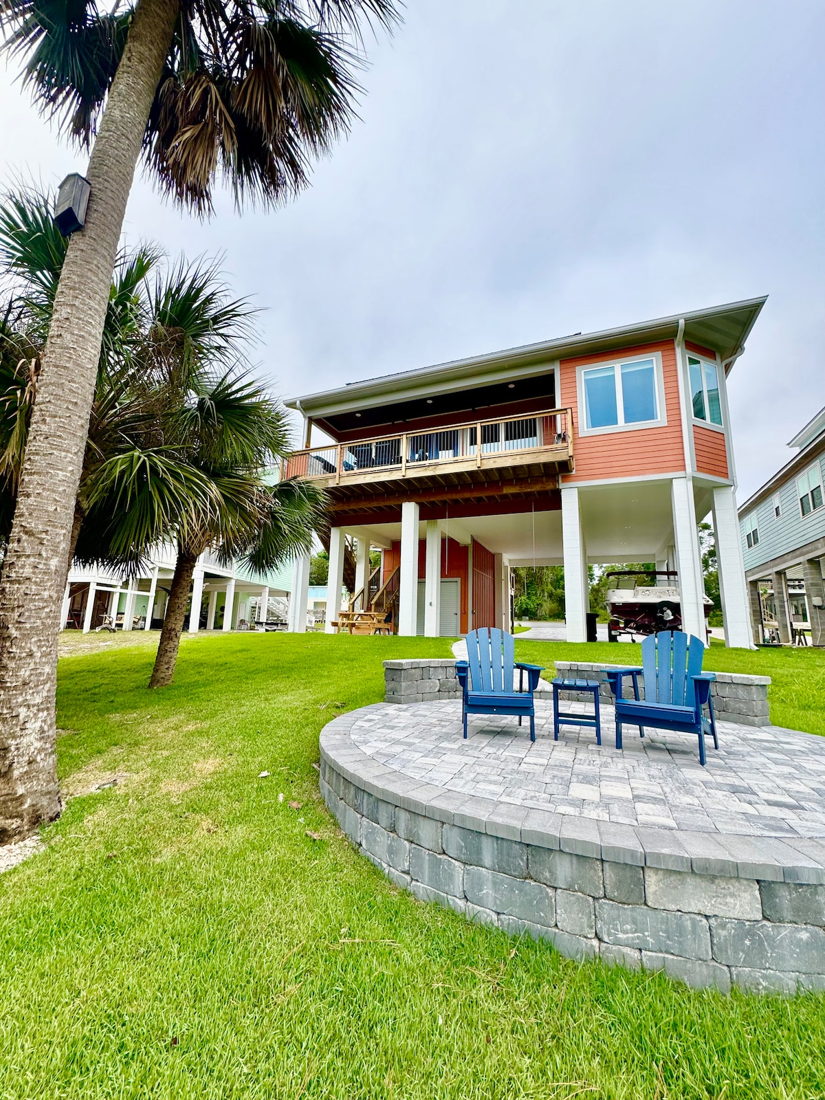 The waterfront home is set on a grassy area, surrounded by palm trees. Underneath the structure, a stone patio features two blue chairs facing a circular stone fire pit, providing a cozy outdoor seating option.