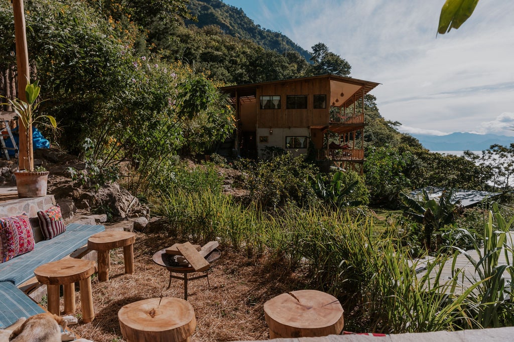 A spacious outdoor area features wooden stools arranged on a grassy patch, surrounded by lush greenery. In the background, a multi-story eco-lodge is situated on the hillside, offering scenic views of the landscape and sky.