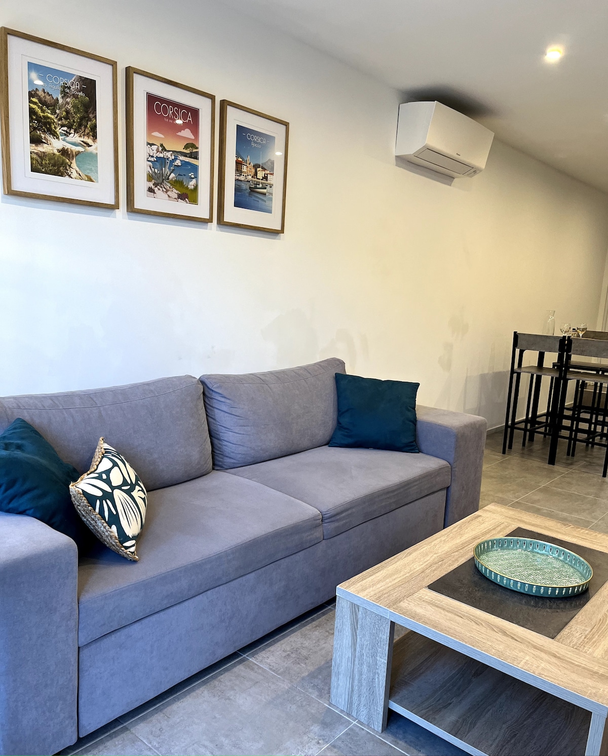 A modern living area features a grey sofa adorned with decorative cushions, positioned near a light wooden coffee table. Framed art pieces depicting Corsican landscapes are displayed on the wall. A dining table with high chairs is visible in the background.
