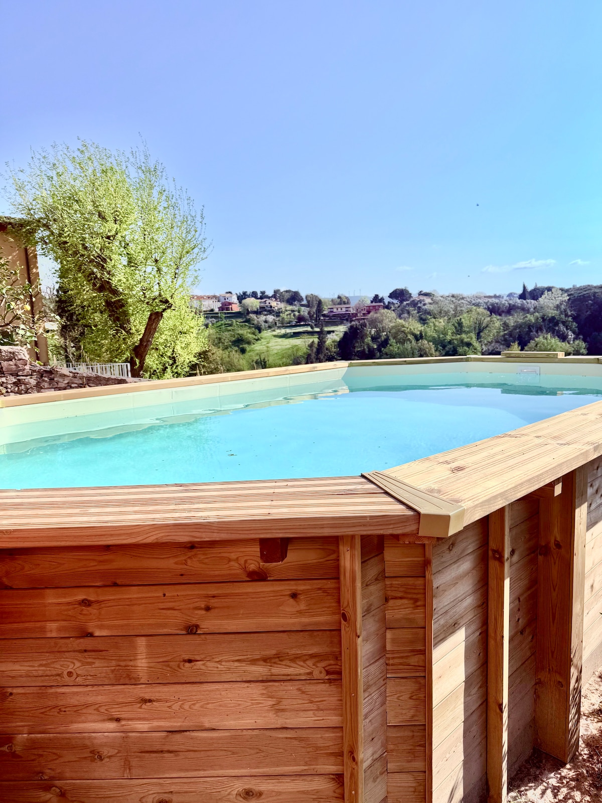 A wooden-framed pool is set against a backdrop of lush hills and clear blue skies. The tranquil water reflects the surroundings, while green trees provide a natural border. Sunlight enhances the inviting atmosphere, making it a serene spot for relaxation.