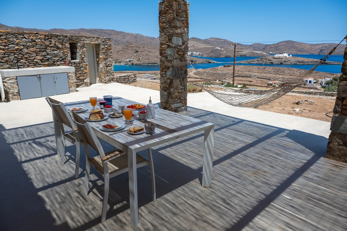 An outdoor dining area features a long table set for breakfast with an array of plates and drinks, surrounded by four white chairs. A hammock hangs nearby, and a stone wall provides partial shade. The view includes a tranquil bay and distant hills.