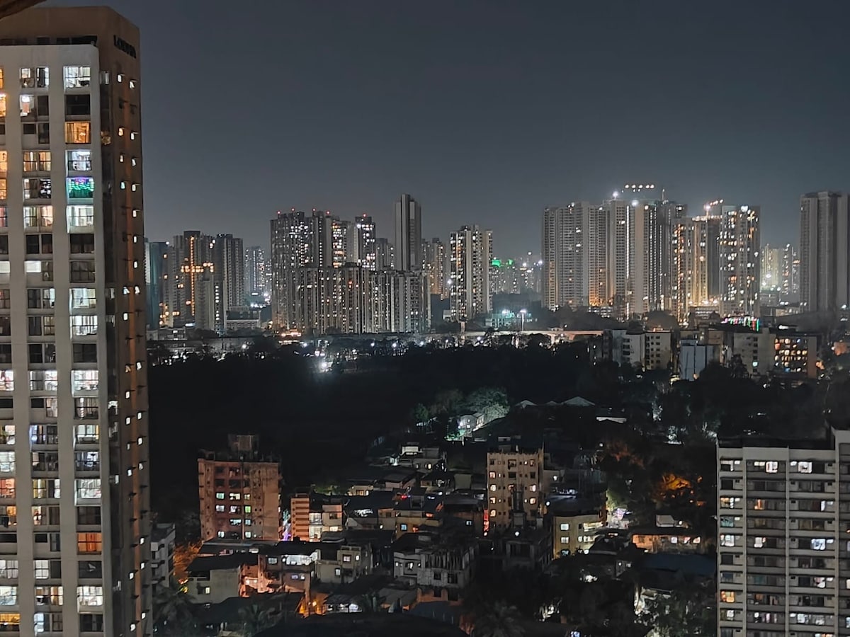 A vibrant city skyline is displayed at night, showcasing numerous illuminated buildings and bustling urban life. The contrast between the bright lights of tall structures and the darker residential areas below creates an immersive view of the city's dynamic atmosphere.