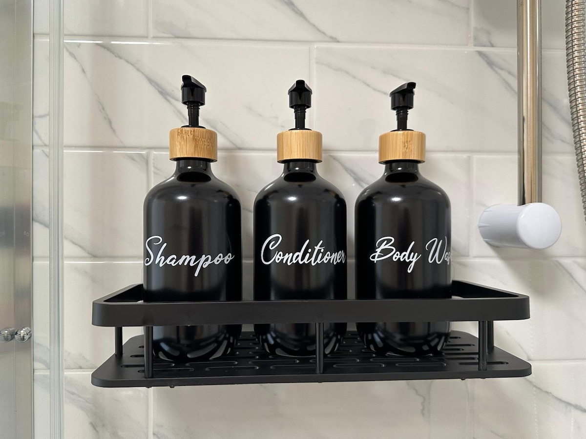A neatly arranged set of three black bottles is displayed on a shelf in the shower. Each bottle features a wooden pump lid, with labels for shampoo, conditioner, and body wash written in white script. The surrounding glass and tile create a clean, modern aesthetic.