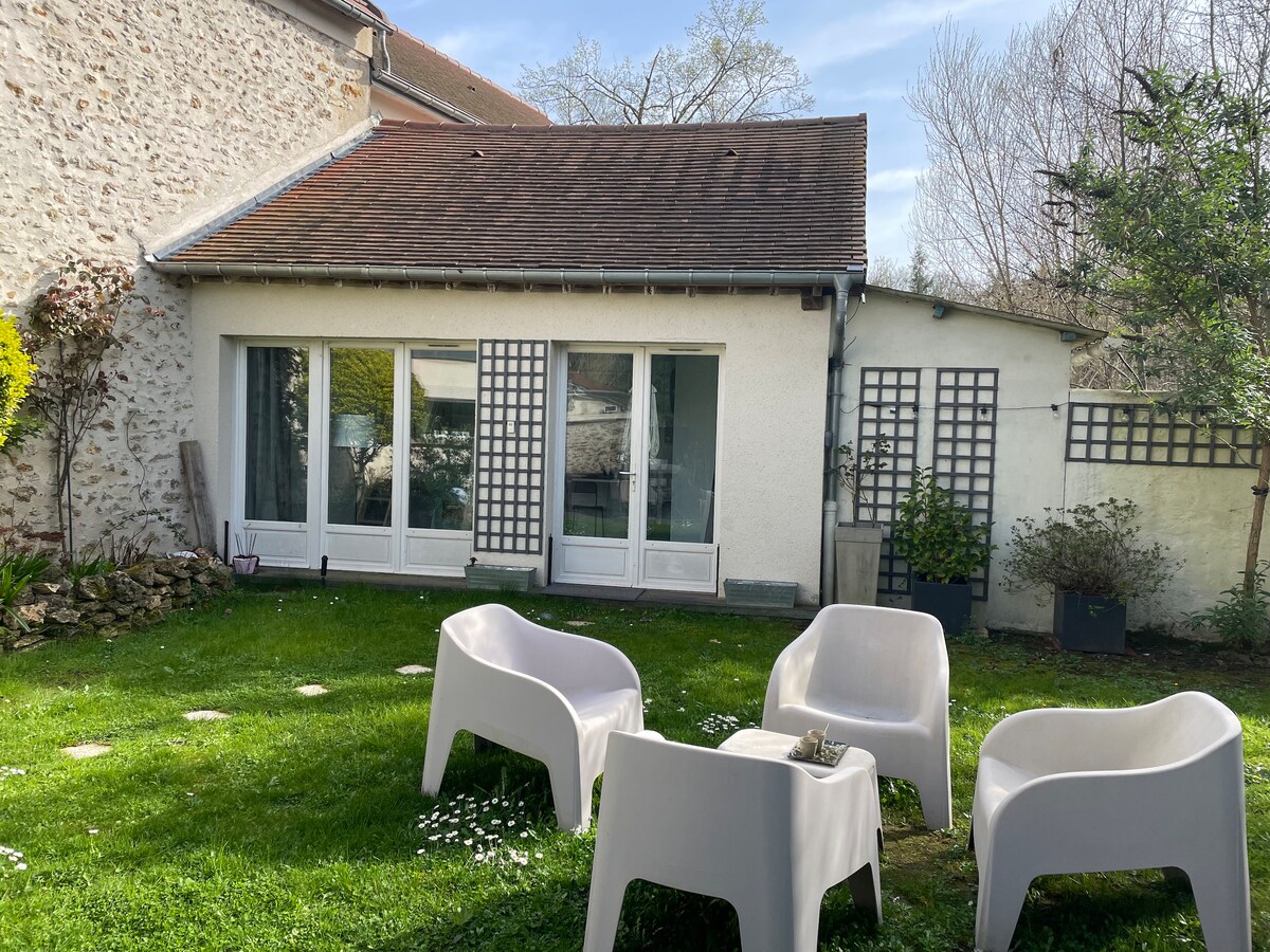 A private garden area features a spacious lawn with green grass and scattered daisies. Light-colored outdoor chairs are arranged around a small table. A wall of large glass doors provides access to a well-lit studio interior, surrounded by landscaped greenery.