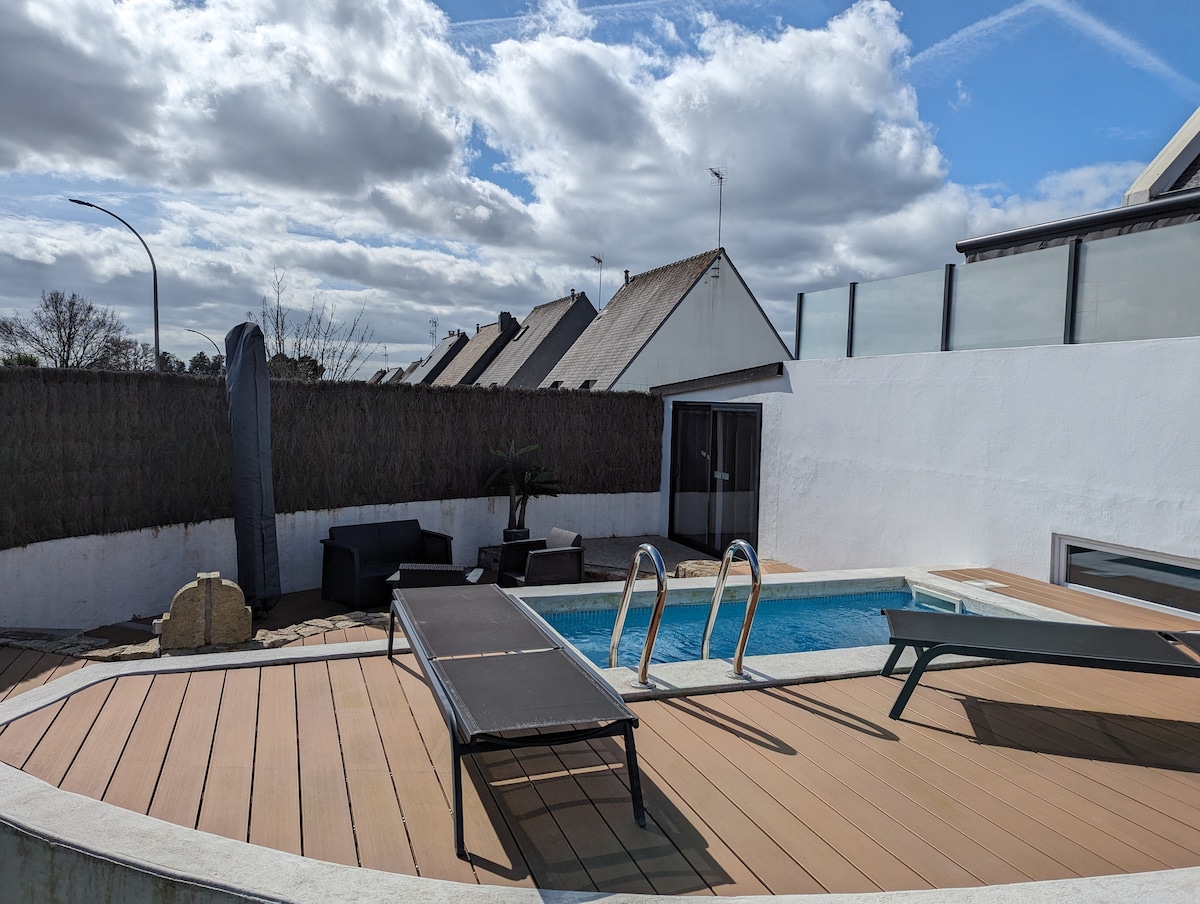 A private outdoor area features a heated swimming pool surrounded by a wooden deck. Sun loungers are positioned near the pool, with a sitting area that includes dark chairs. A low stone structure and greenery provide subtle decoration, complemented by a clear sky with scattered clouds.