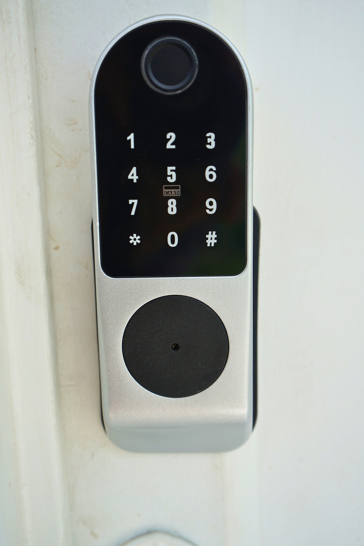 A digital keypad lock is mounted on a white door. The keypad features numbers 0 through 9, along with symbols for enter and clear. The sleek design includes a round keyhole at the bottom, providing a modern touch to the entrance.