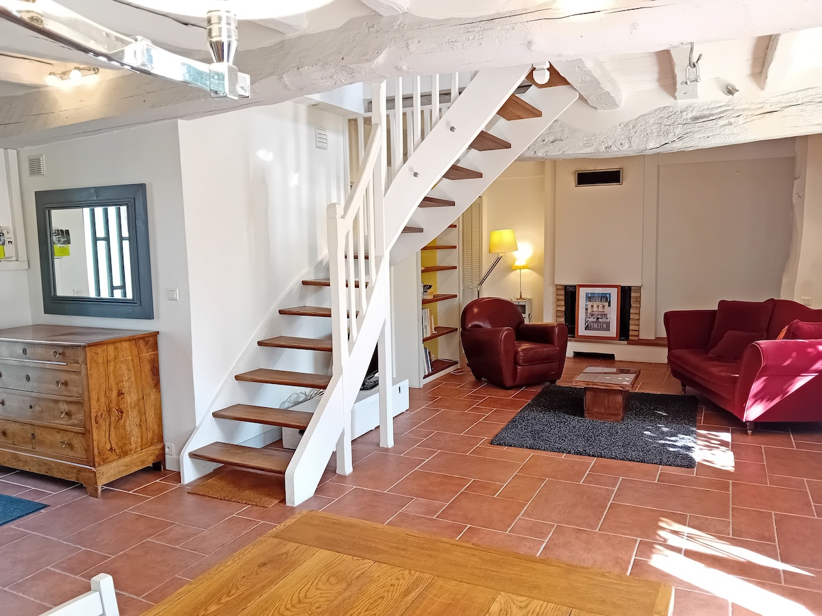 A bright and spacious living area features a wooden dining table and a cozy seating arrangement with two red armchairs and a sofa. Natural light floods the room, highlighting the warm terracotta flooring and whitewashed beams. A staircase leads to the upper level, enhancing the open layout.